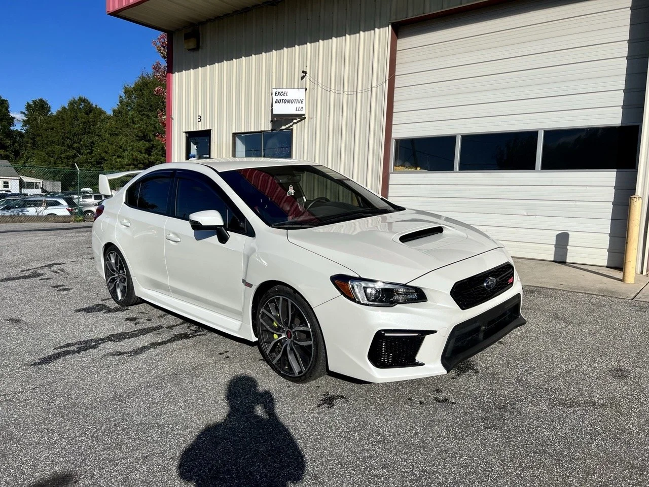 A white Subaru WRX STI parked outside a building with a sign for Excel Automotive LLC. The car has a hood scoop, tinted windows, and black wheels, with a small rear spoiler.