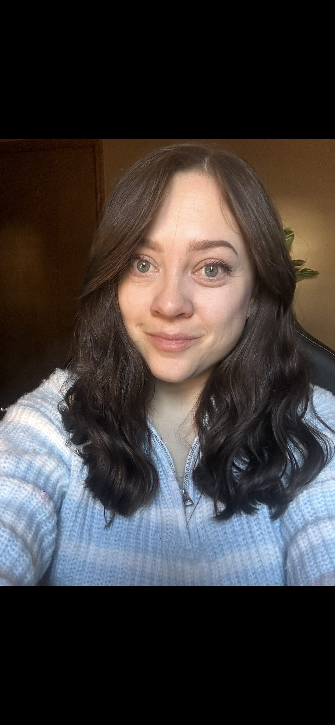 A woman with long, wavy dark hair and blue eyes taking a selfie indoors, smiling softly, wearing a light blue zip-up sweater.