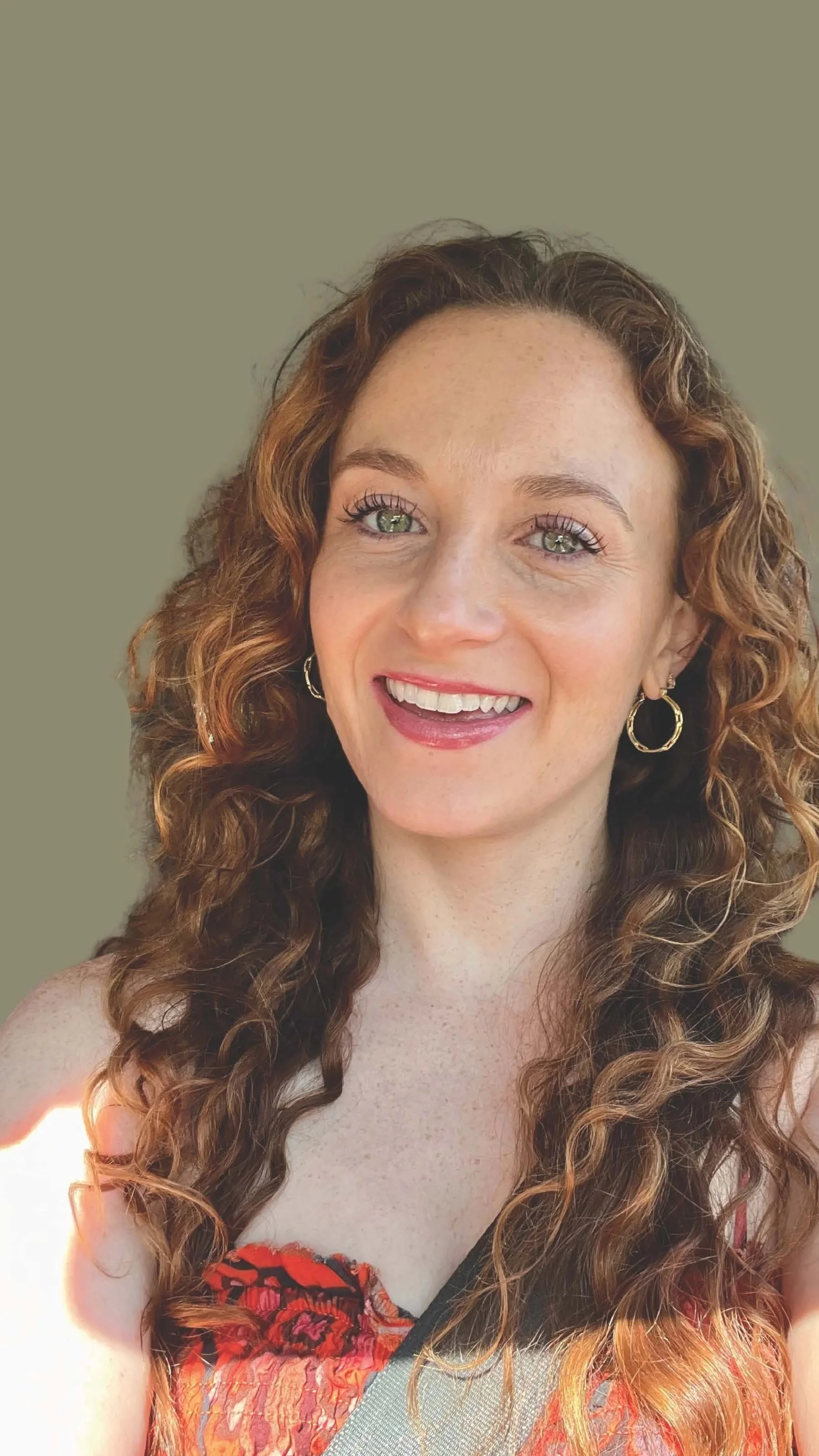 A woman with long, curly red hair, green eyes, and freckles, smiling for a selfie, wearing hoop earrings and a colorful top, with a plain background.