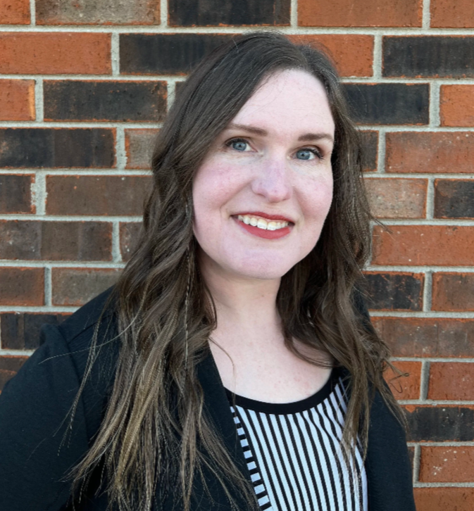 A woman with long brown wavy hair and blue eyes smiling in front of a brick wall.