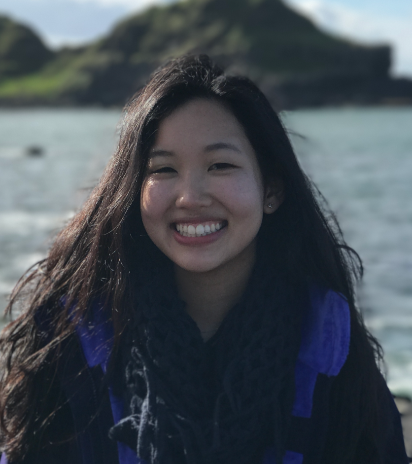 Smiling young woman with long black hair, standing outdoors near water with a hilly landscape in the background.