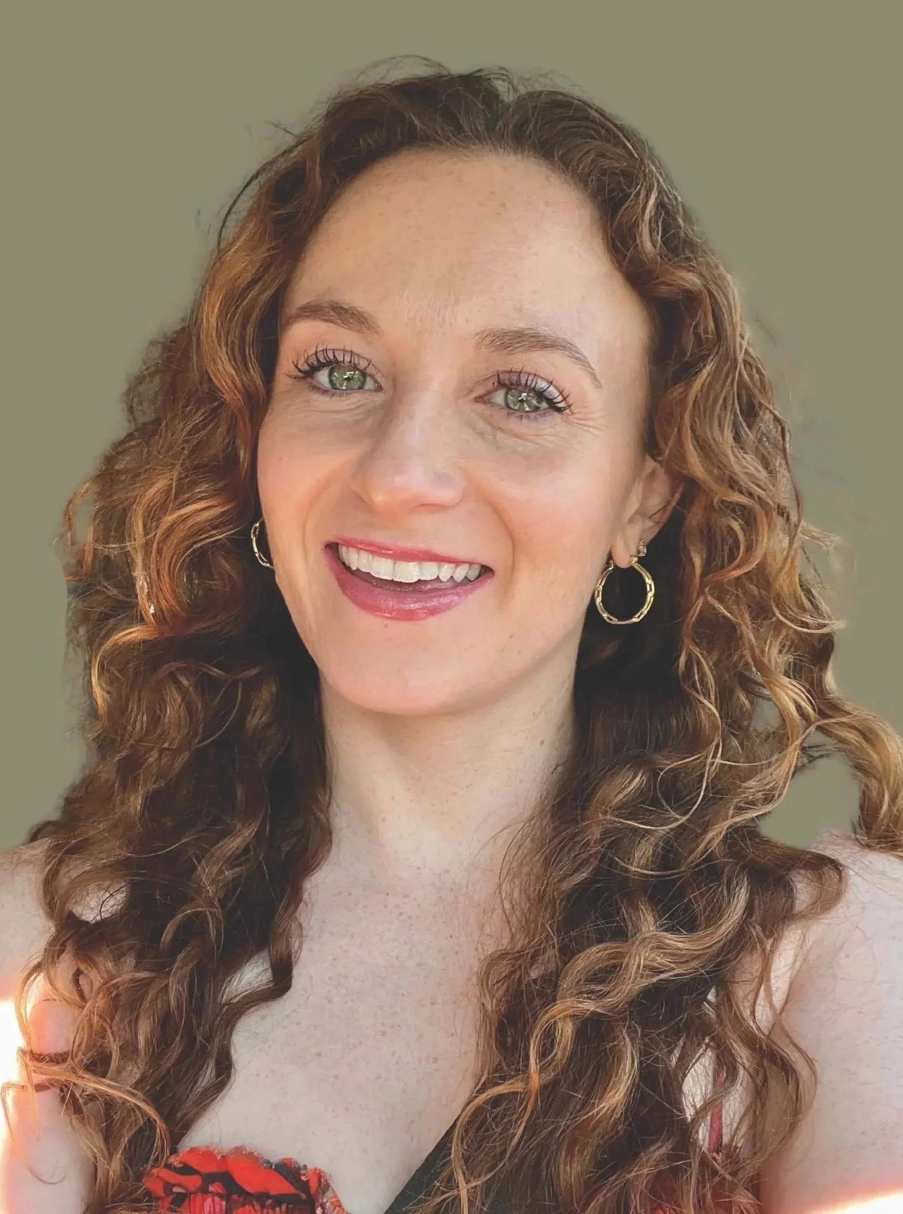A young woman with curly red hair, green eyes, and a bright smile, wearing hoop earrings and a floral top, shouldering a friendly expression with a neutral background.