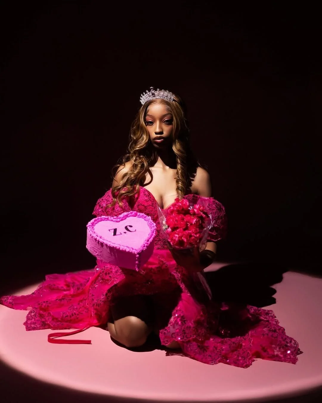 A young woman in a pink lace dress with long, wavy hair, wearing a crown, kneeling on a pink surface in a dark background. She holds a heart-shaped box of chocolates labeled '3. Z' and a bouquet of pink roses.