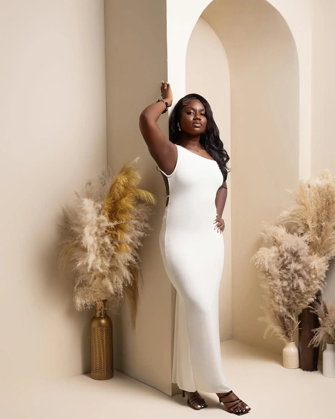 A woman in a white sleeveless gown posing with her hand on her hip in a minimalist beige room with decorative pampas grass in vases.