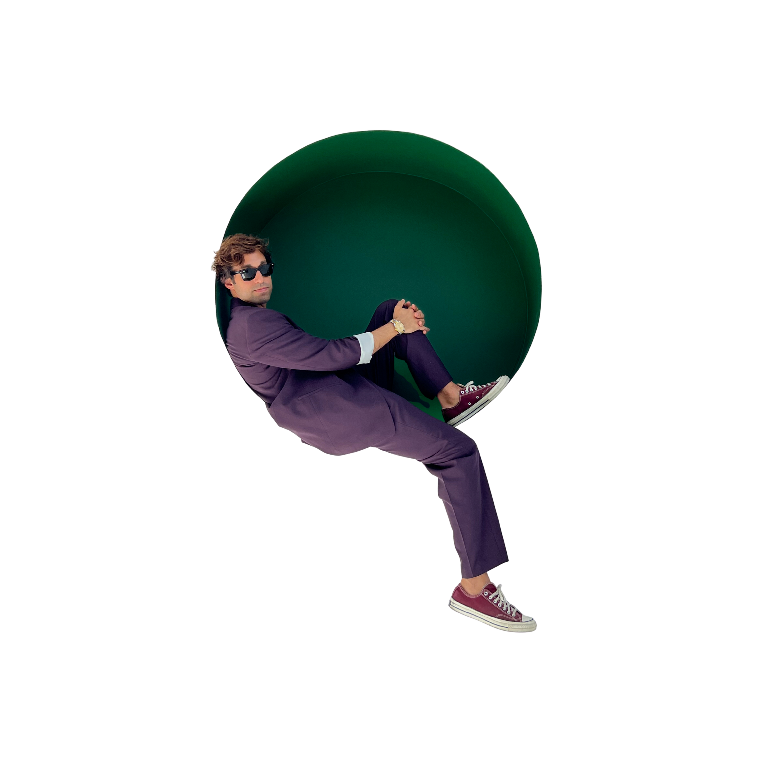 A man wearing sunglasses, a navy blazer, and purple pants sitting in a green oversized pipe with purple sneakers.