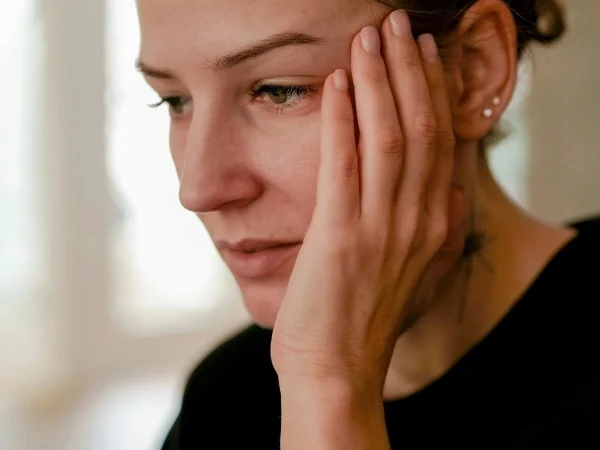 Close-up of a woman with brown hair, wearing a black shirt, touching her face with her right hand, looking down thoughtfully.