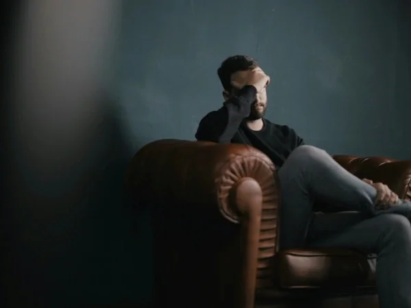 A man sitting on a leather couch, resting his head in his hand, appearing distressed or deep in thought, in a dimly lit room with a dark wall background.