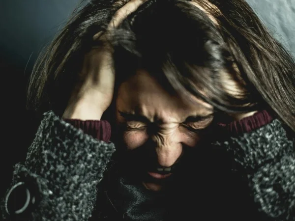 A woman with long brown hair holds her head with both hands, her face showing distress or pain, wearing a patterned coat and a maroon shirt underneath.