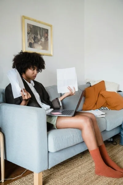 Woman sitting on couch with computer on lap holding papers in each hand looking confused