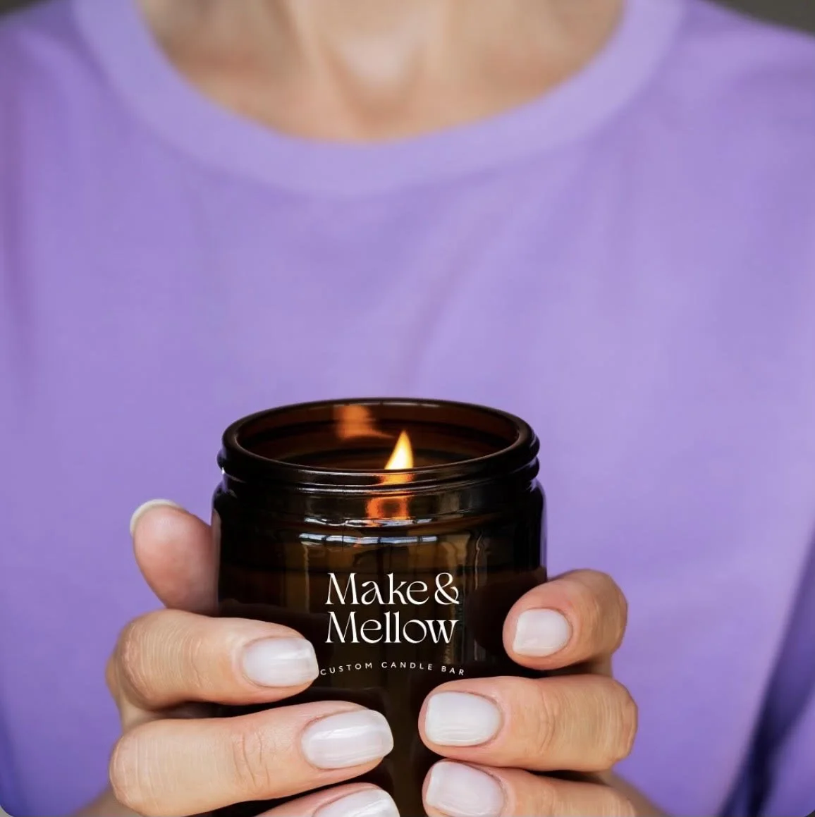 Person holding a lit candle in a dark amber jar labeled "Make & Mellow" with a purple shirt background.