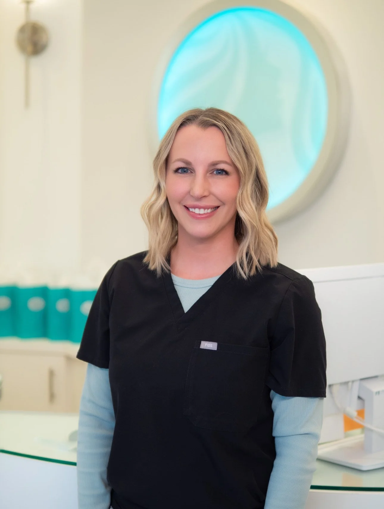 A smiling woman in medical scrubs standing in a clinical setting with a round, glowing blue art piece on the wall behind her.