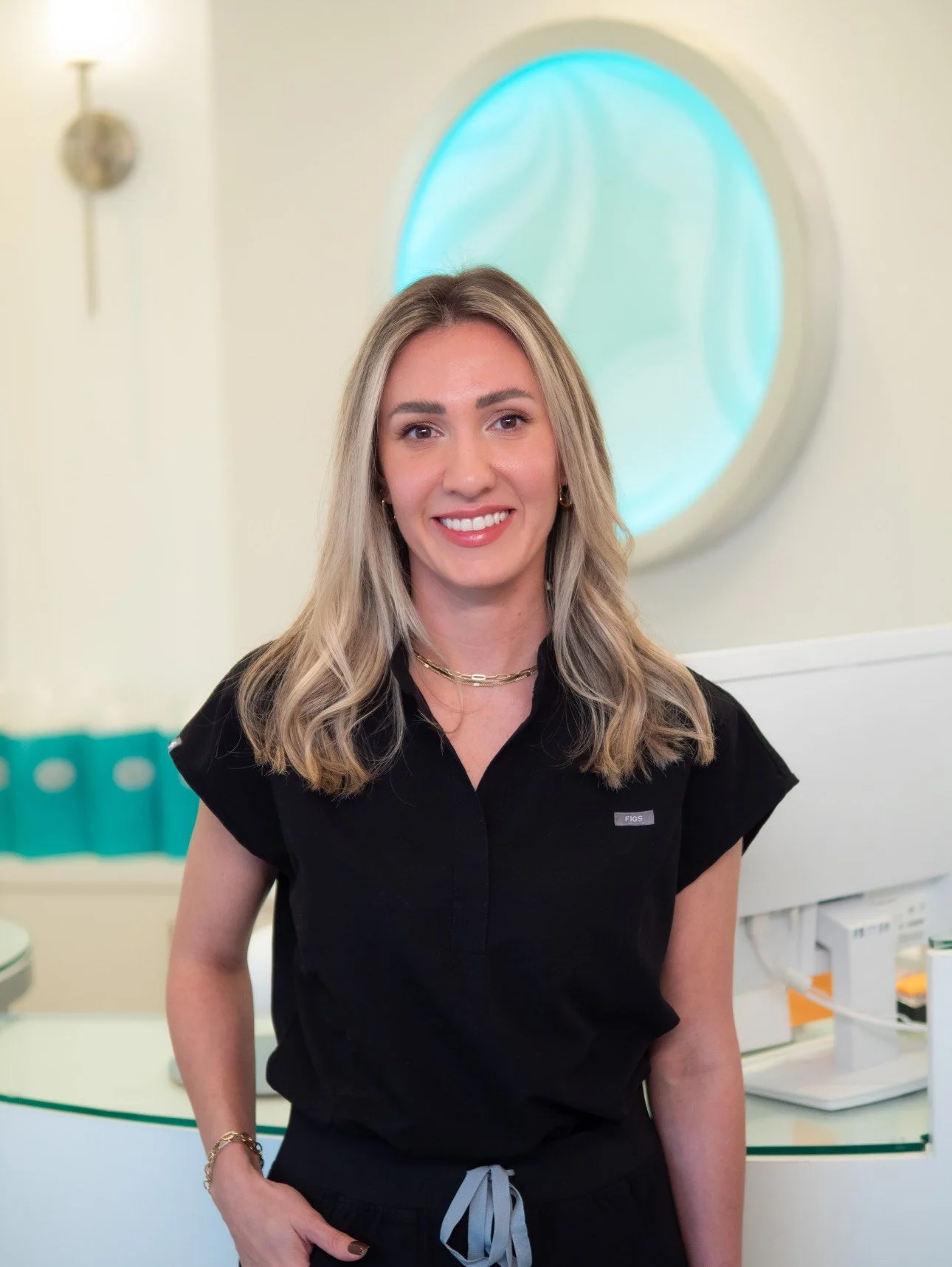 A woman with blonde hair wearing a black top and dark pants stands in a bright, modern medical or dental office, smiling at the camera.