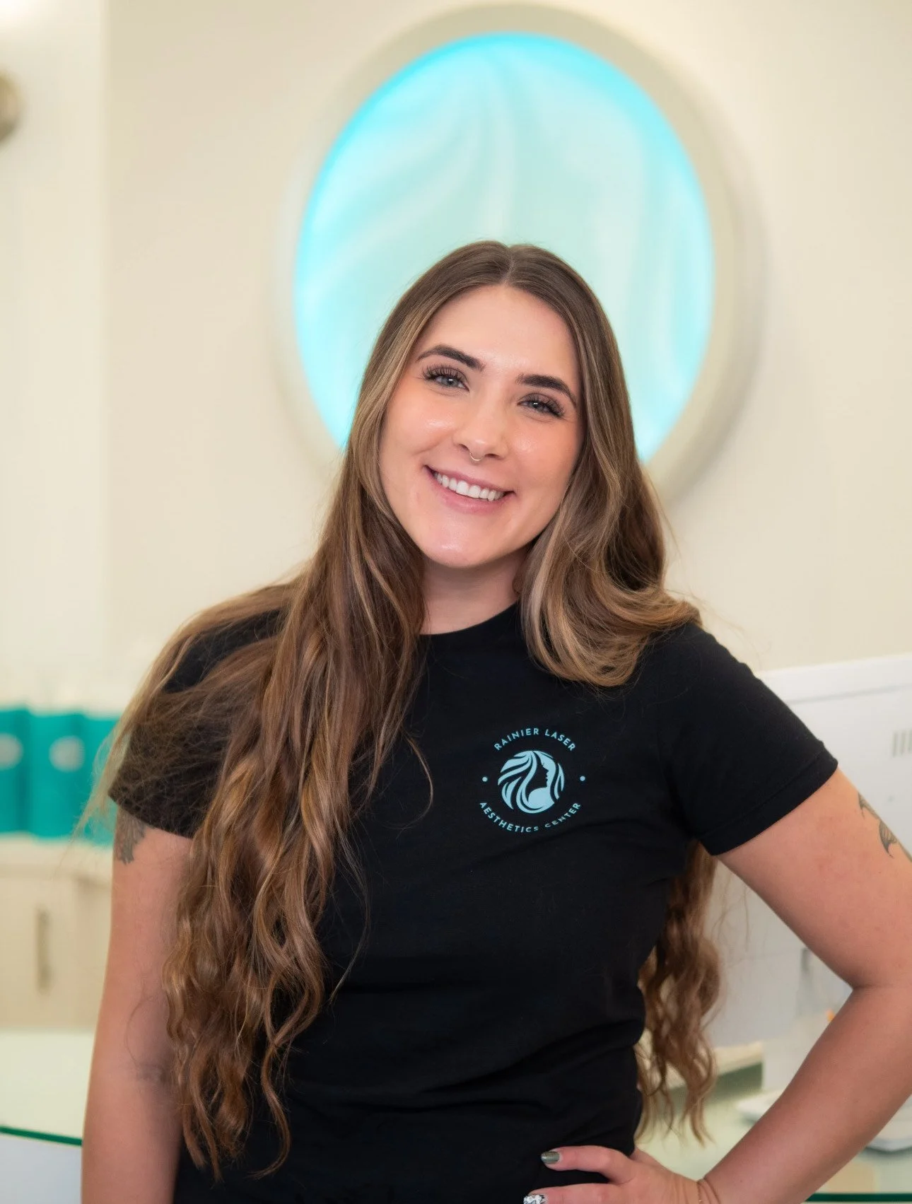 A smiling woman with long wavy brown hair, wearing a black t-shirt with a logo that reads 'Rainier Laser Aesthetic Center,' standing indoors in front of a light-colored wall with a blue-lit circular decoration.