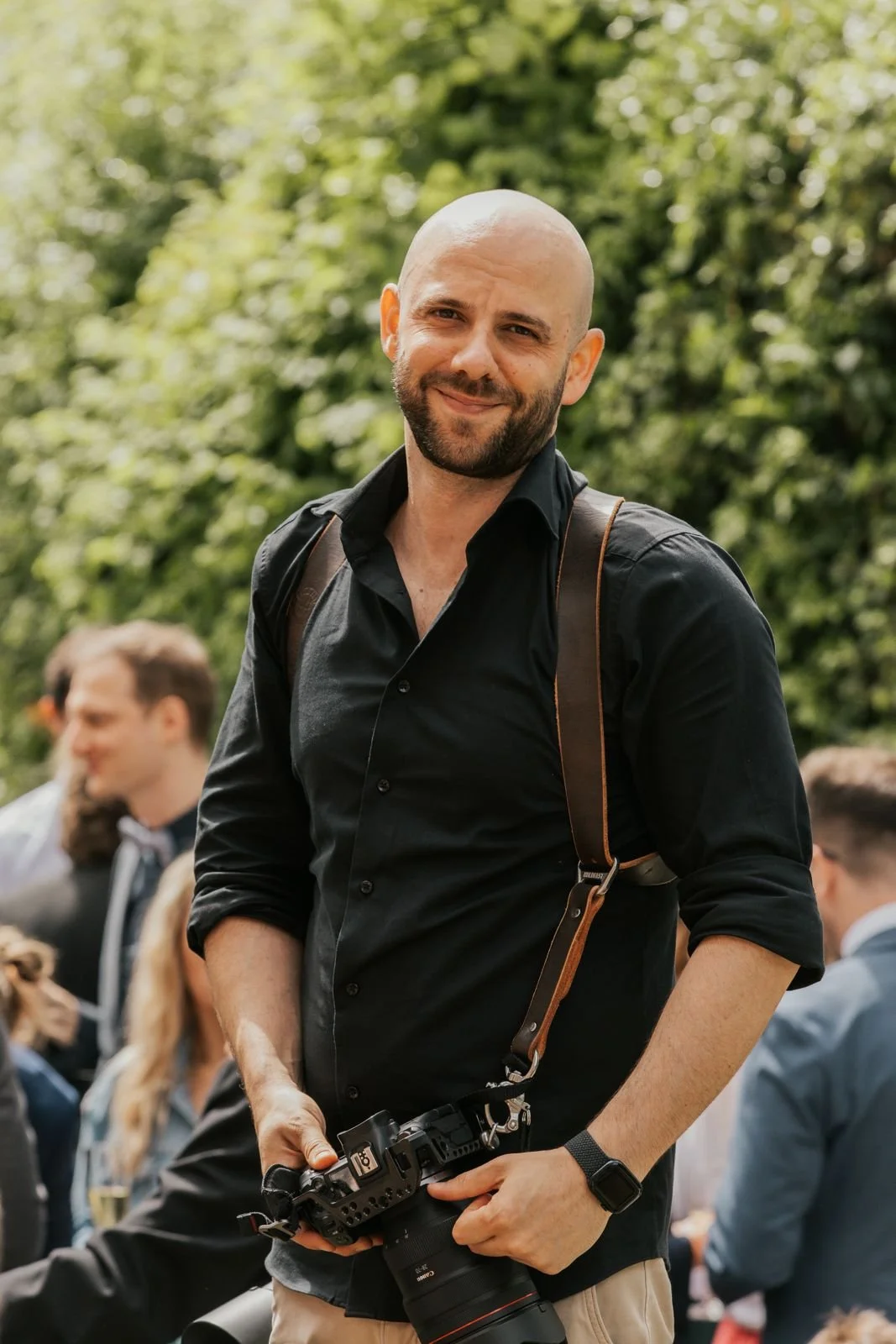 Ein lächelnder Mann mit einem schwarzen Hemd und einer Kamera in der Hand, im Freien bei einer Veranstaltung, umgeben von anderen Menschen im Hintergrund.
