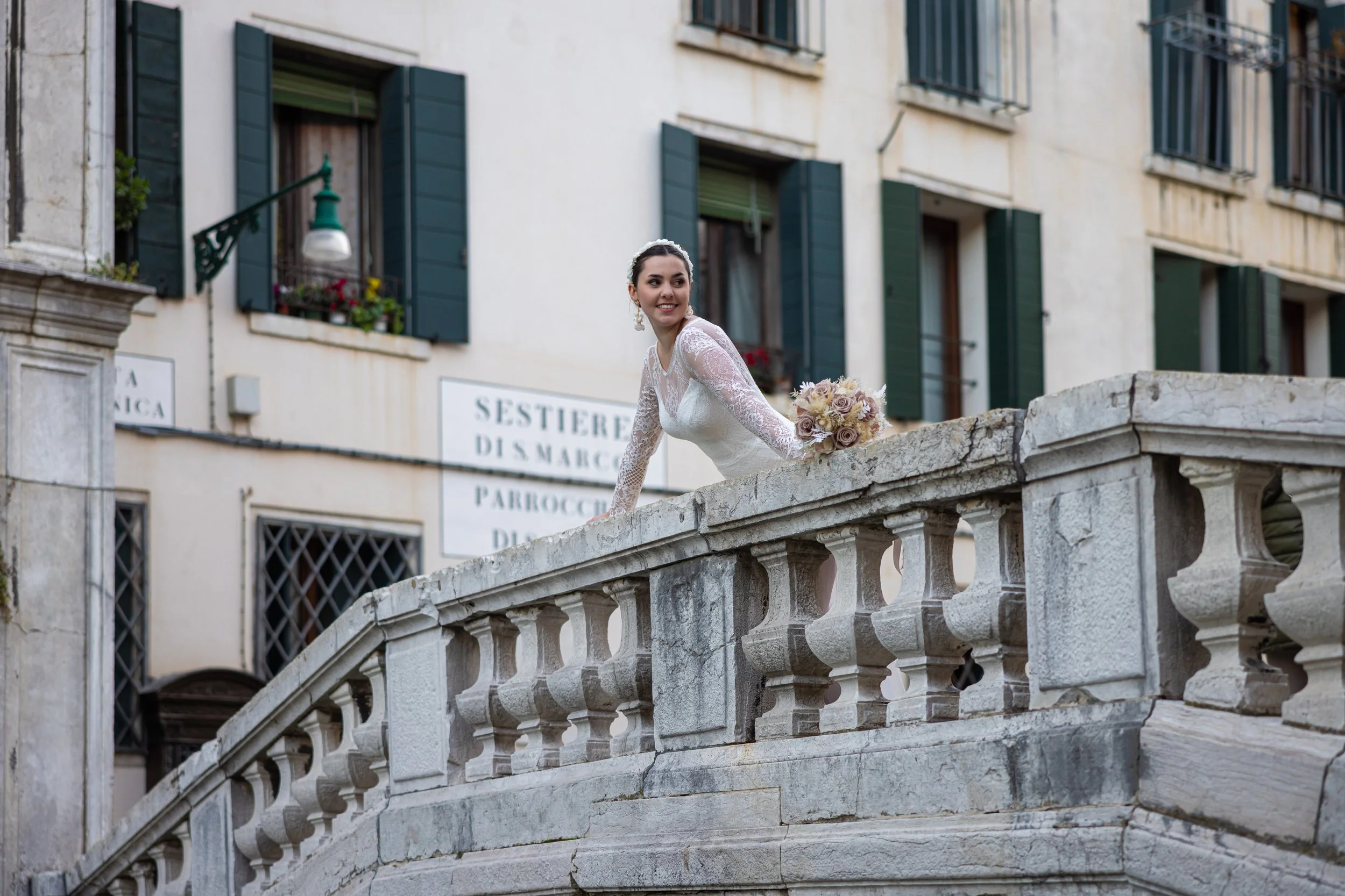 Braut in weißem Brautkleid mit Blumenstrauß steht auf einer Brücke in Venedig.
