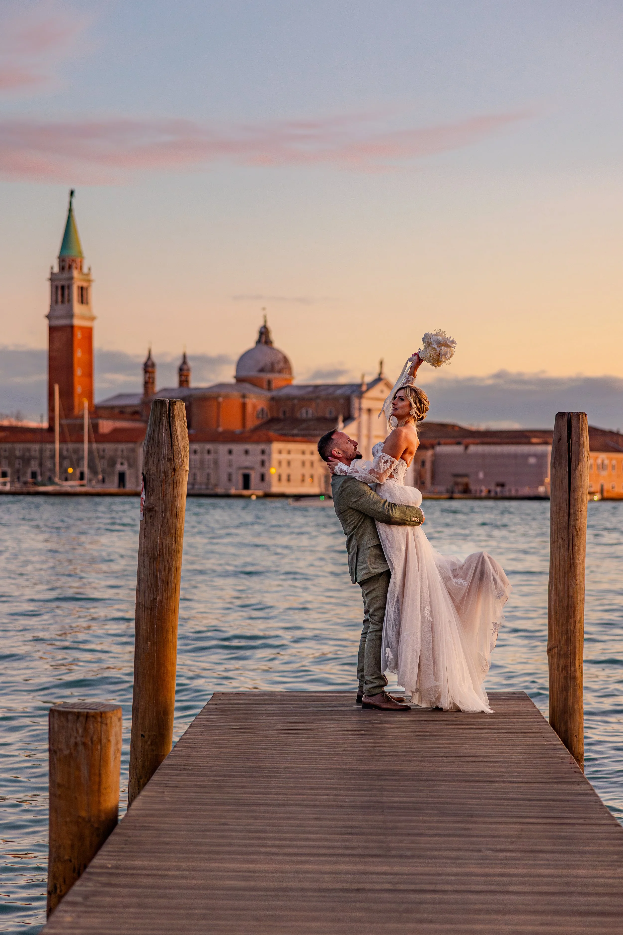 Ein Hochzeitspaar auf einem Steg am Wasser, mit Blick auf eine Stadt mit Kirchtürmen, bei Sonnenuntergang.