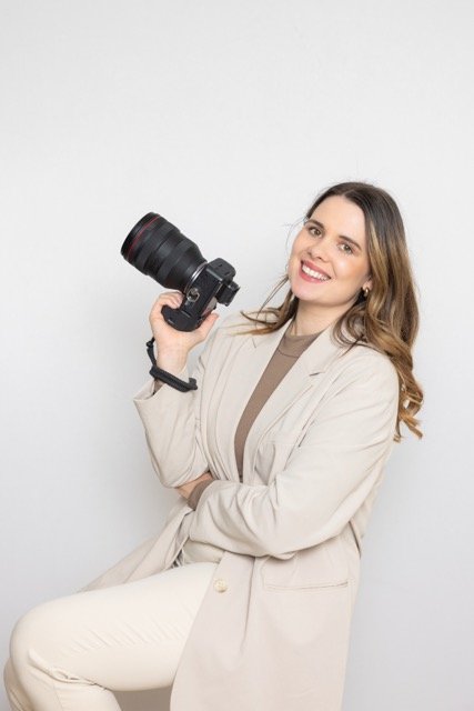 Junge Frau in beige Jacke hält eine professionelle Kamera mit großem Objektiv, lächelt und sitzt vor weißem Hintergrund.