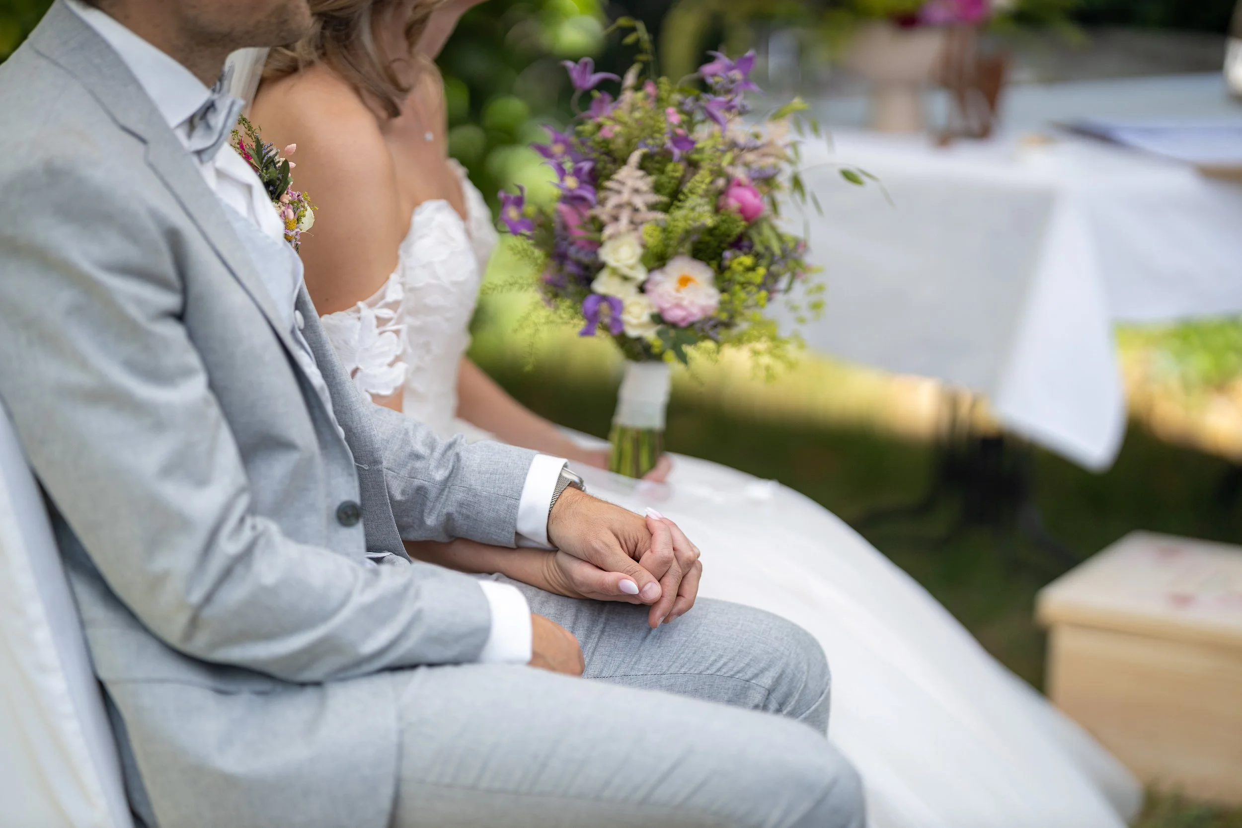 Ein Brautpaar sitzt bei ihrer Hochzeit, hält Händchen, im Hintergrund ist ein Blumenstrauß sichtbar.