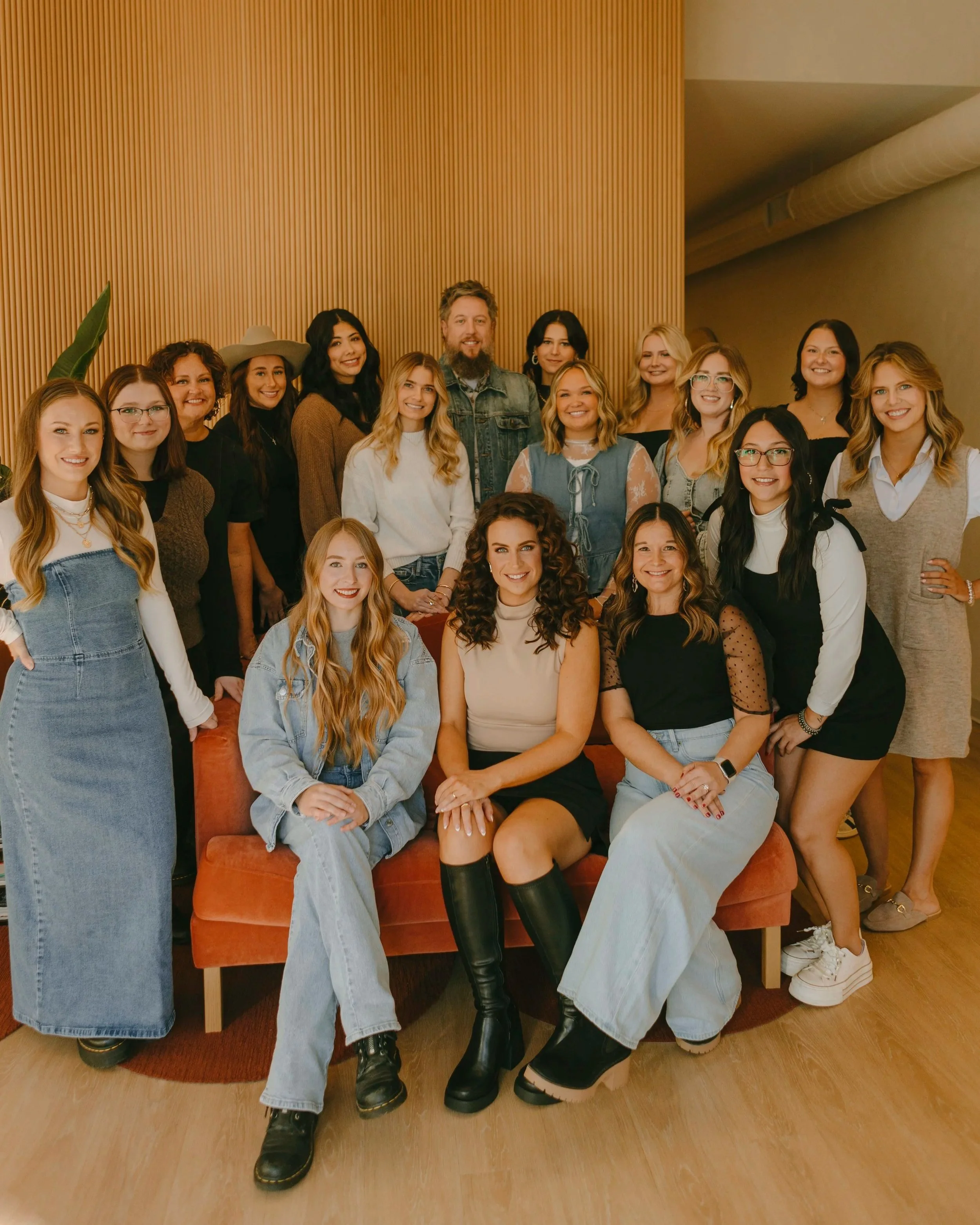 Group of 17 women and one man posing together indoors. They are standing and sitting in front of a wooden wall, smiling at the camera. Salon branding session.