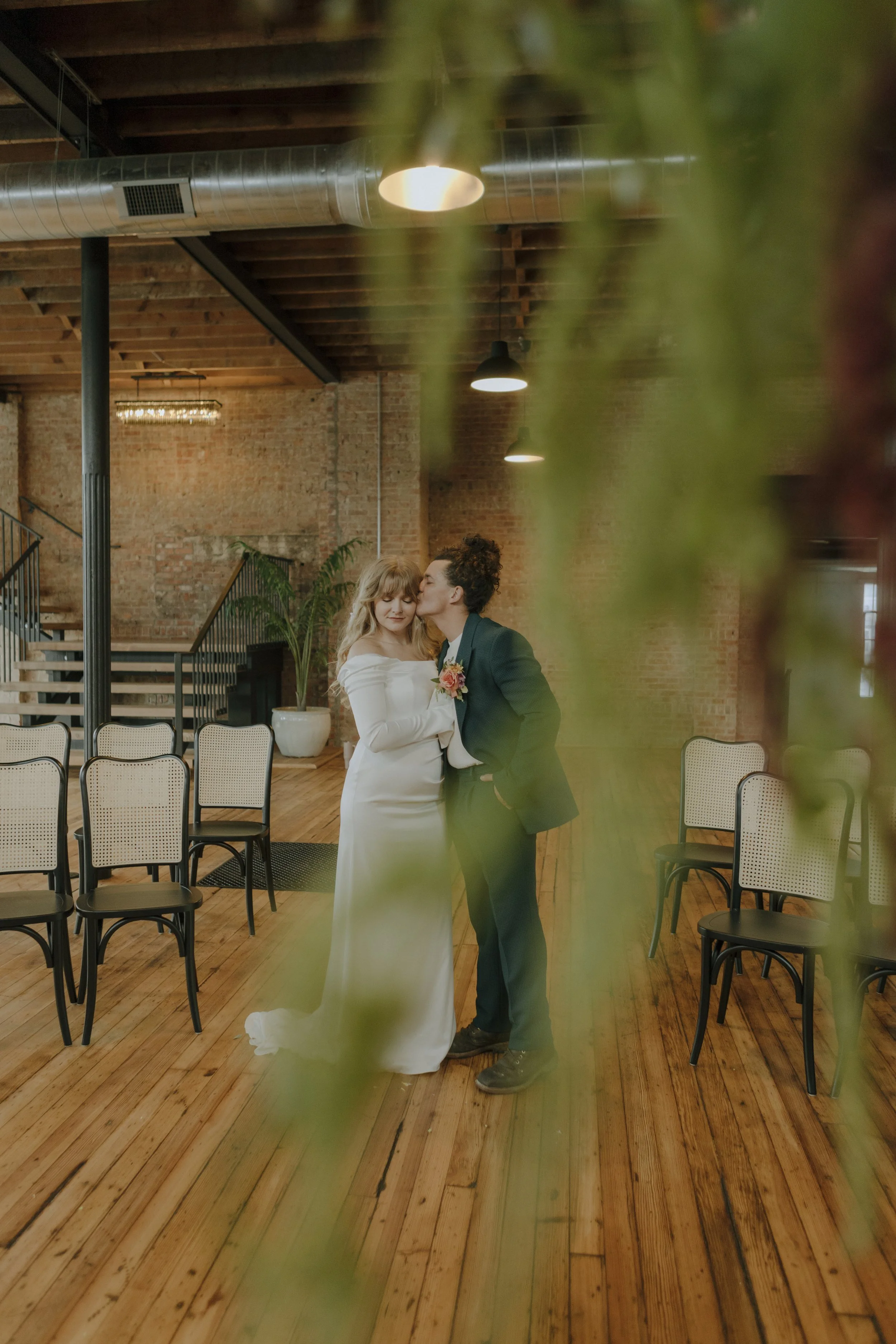 A bride and groom sharing a kiss in a rustic indoor wedding venue with brick walls and wooden floors, surrounded by empty chairs.