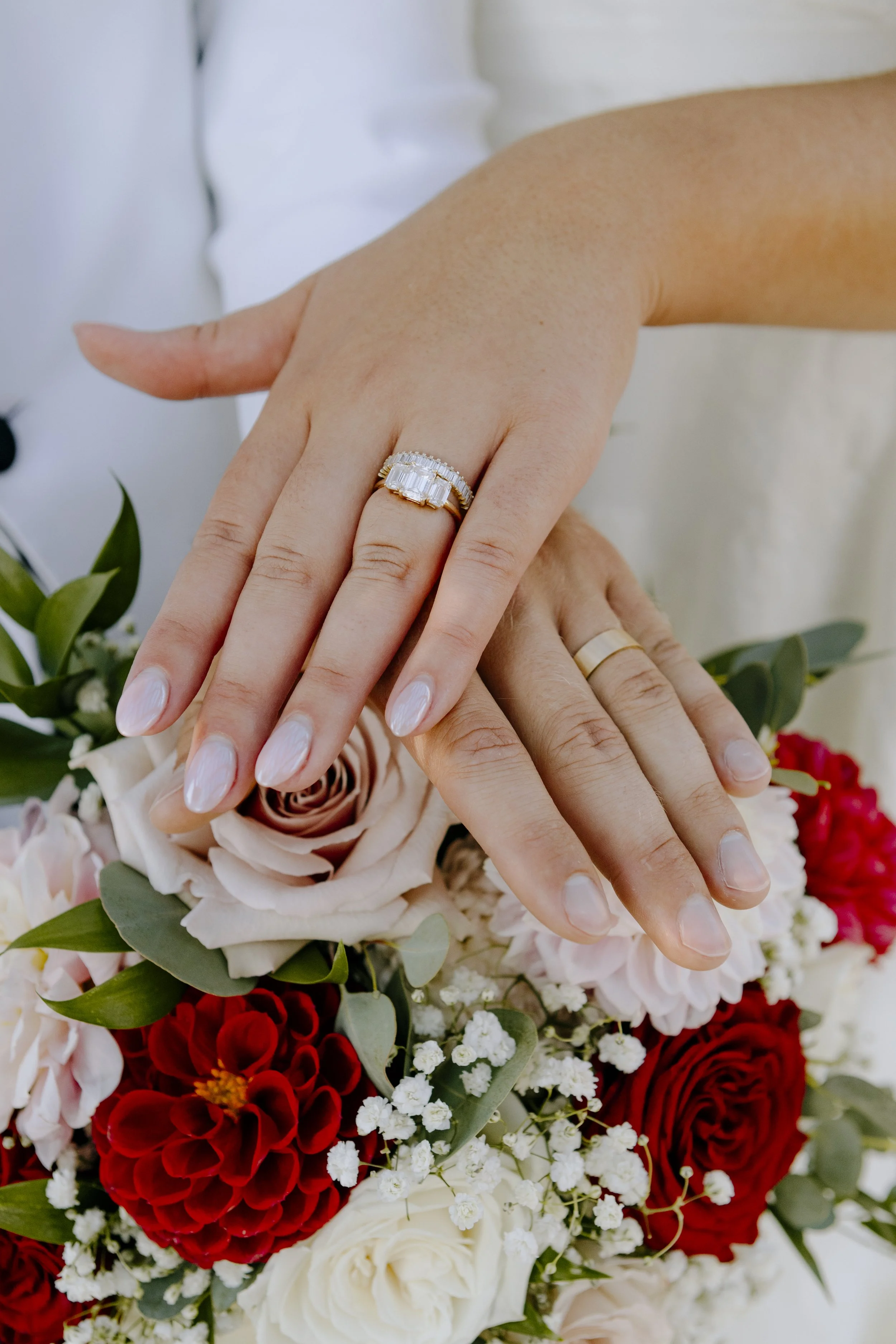 Close-up of a person's hands with wedding rings, resting on a bouquet of red, white, and pink flowers.