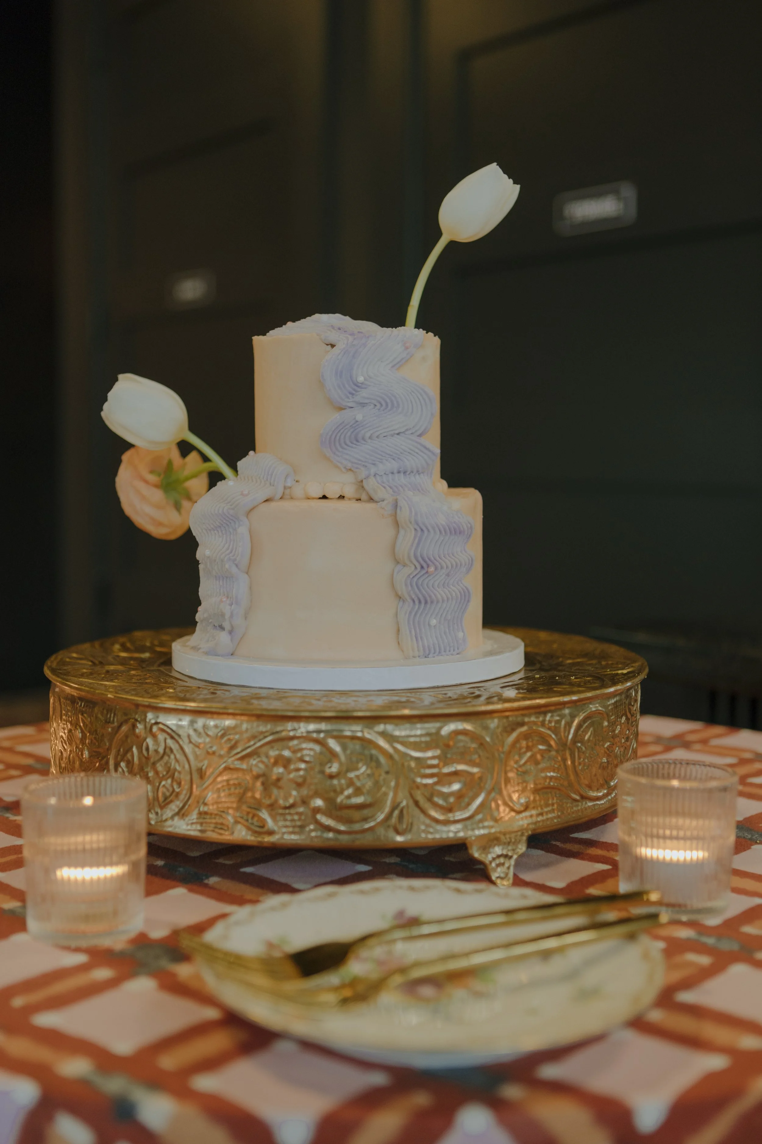 A two-tiered wedding cake with white and lavender frosting, decorated with white tulip flowers, on an ornate gold stand, surrounded by lit candles and a dessert plate with a gold fork, set on a checkered tablecloth.