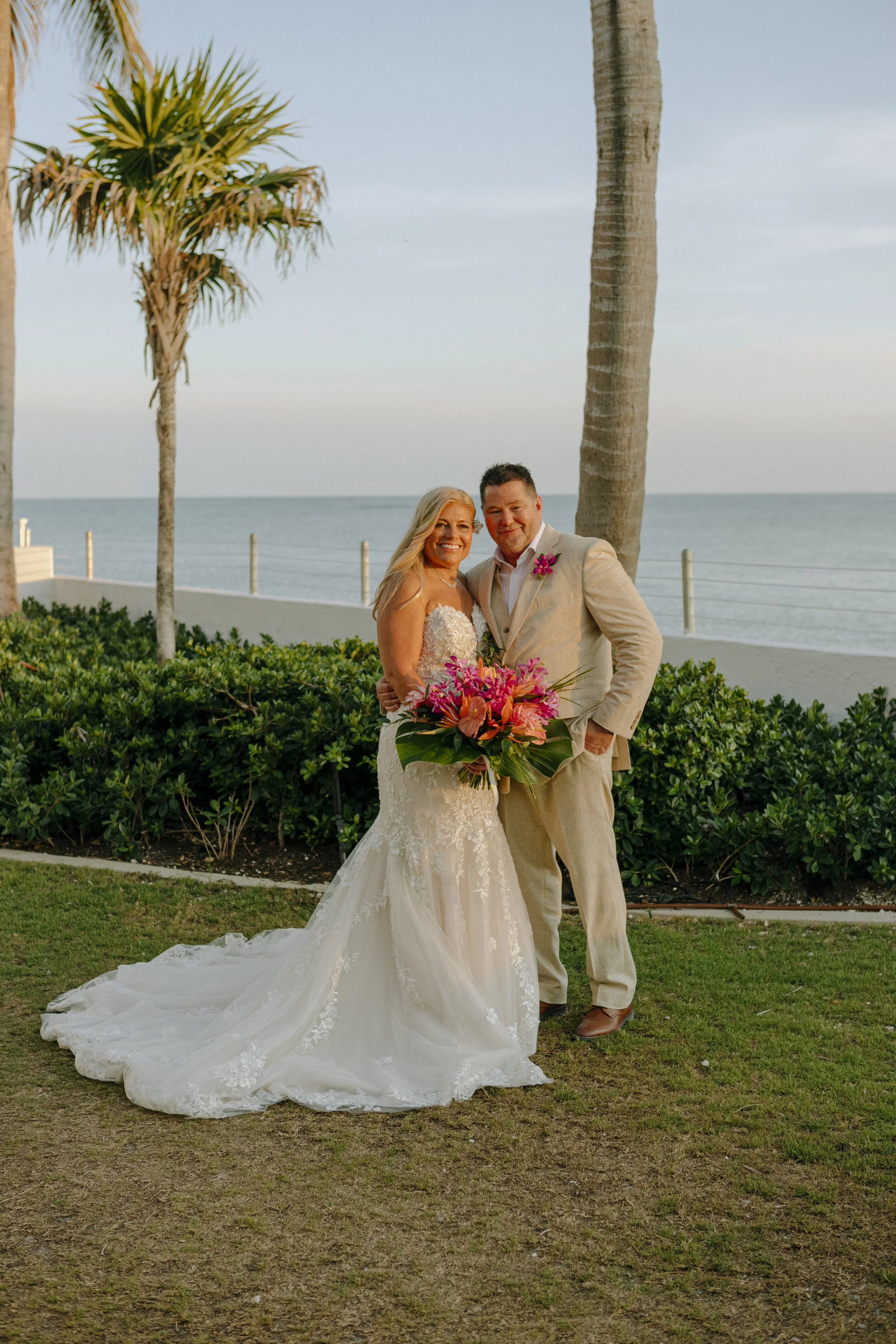 A bride in a white lace wedding gown holding a pink and orange floral bouquet, standing next to a groom in a light-colored suit with a pink floral boutonniere, outdoors near the beach with palm trees and ocean in the background at sunset.
