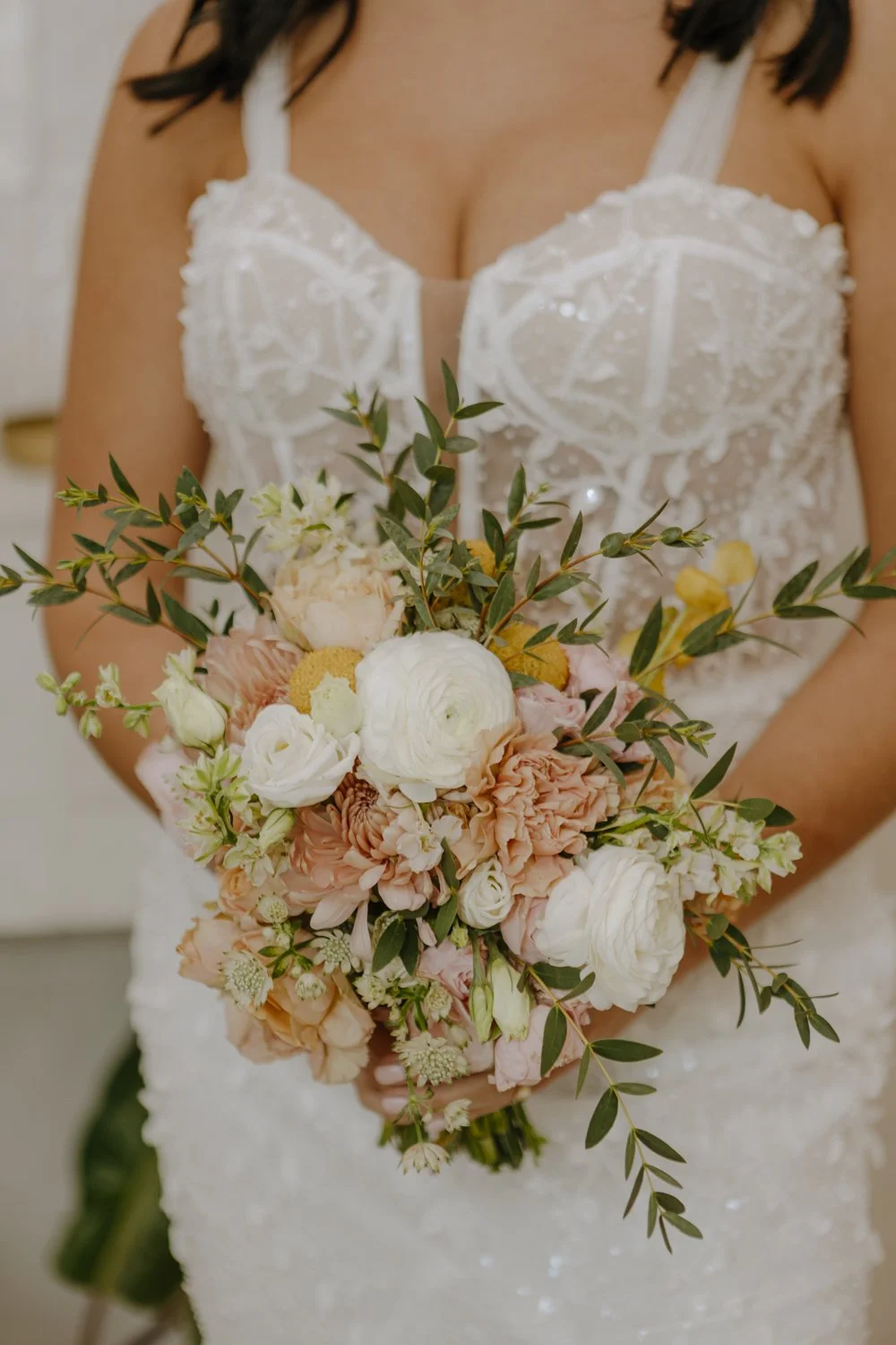 A bride holding a bouquet of white, peach, and yellow flowers with green foliage, in front of her white lace wedding dress.