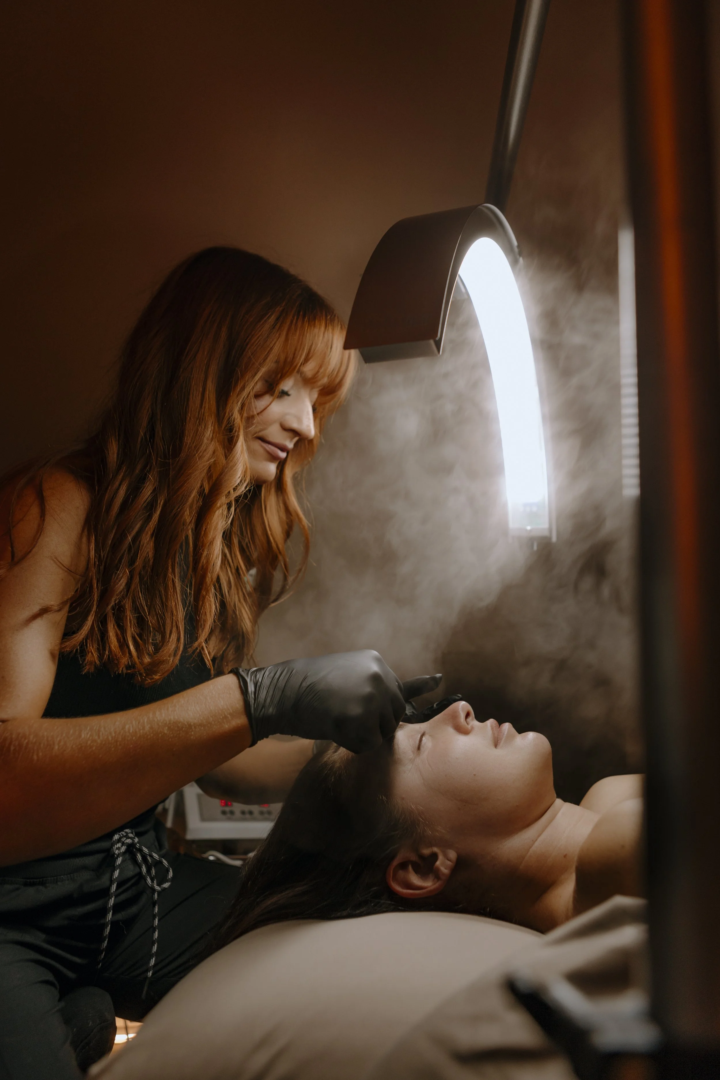 A woman lying on a bed with her eyes closed, getting a cosmetic or dermatological treatment on her eyebrows from a professional wearing black gloves.