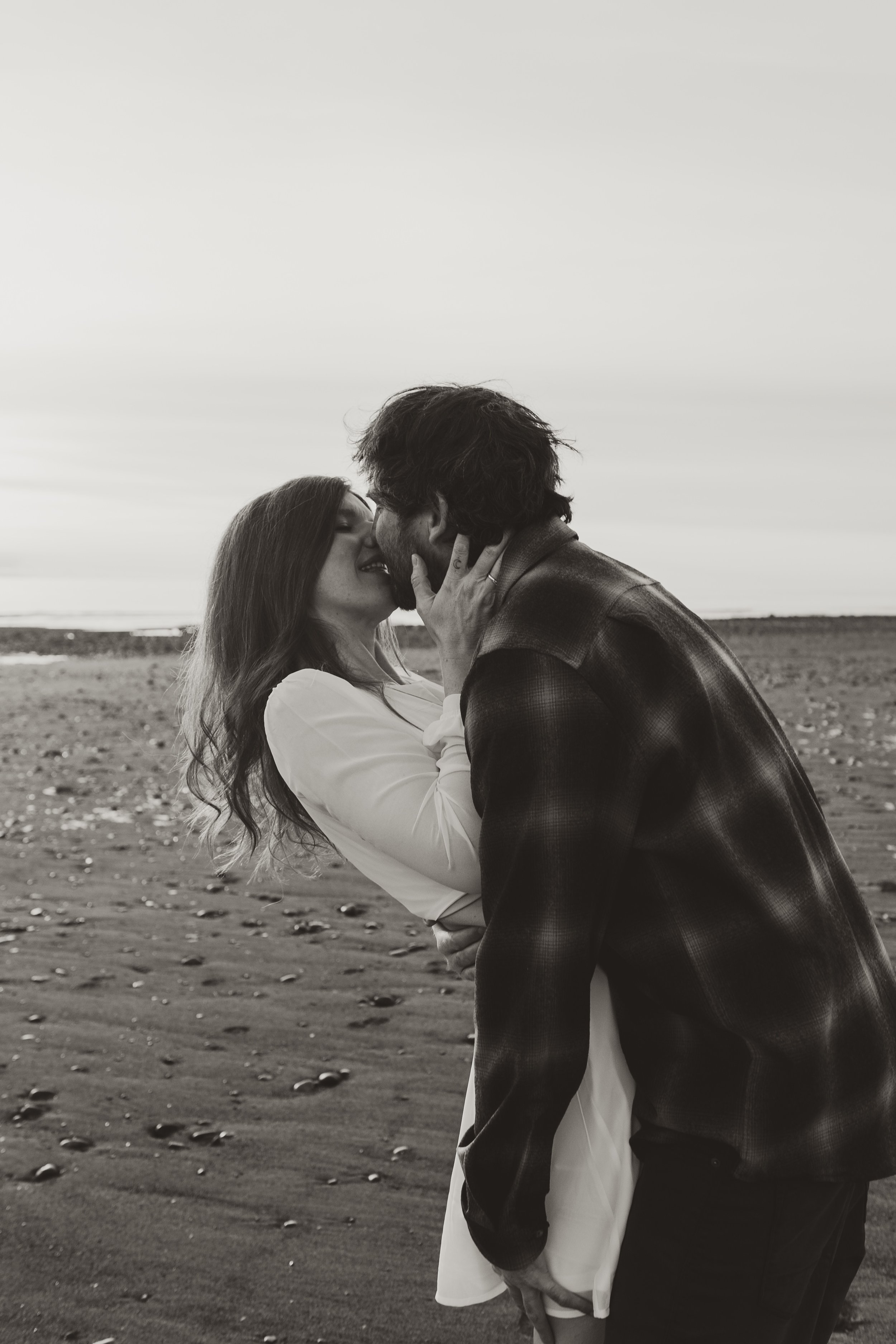 A black and white photo of a couple kissing on the beach, with the woman holding the man's face and the man leaning in towards her.