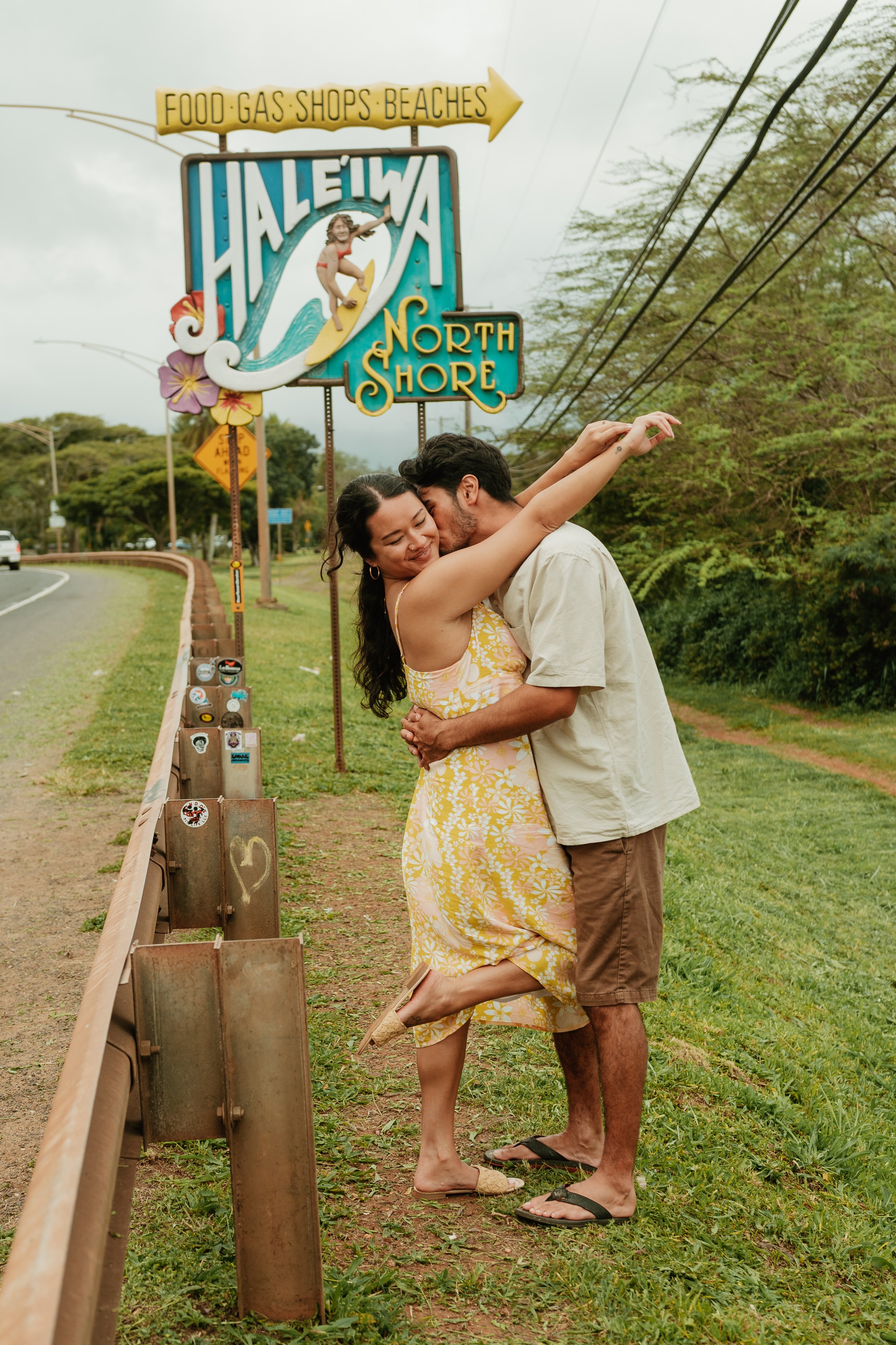 A couple embracing and kissing on the side of a road near a Hawaii sign that says 'Haleiwa North Shore'. The woman is wearing a yellow floral dress with sandals, and the man is in a beige shirt and shorts with flip-flops.