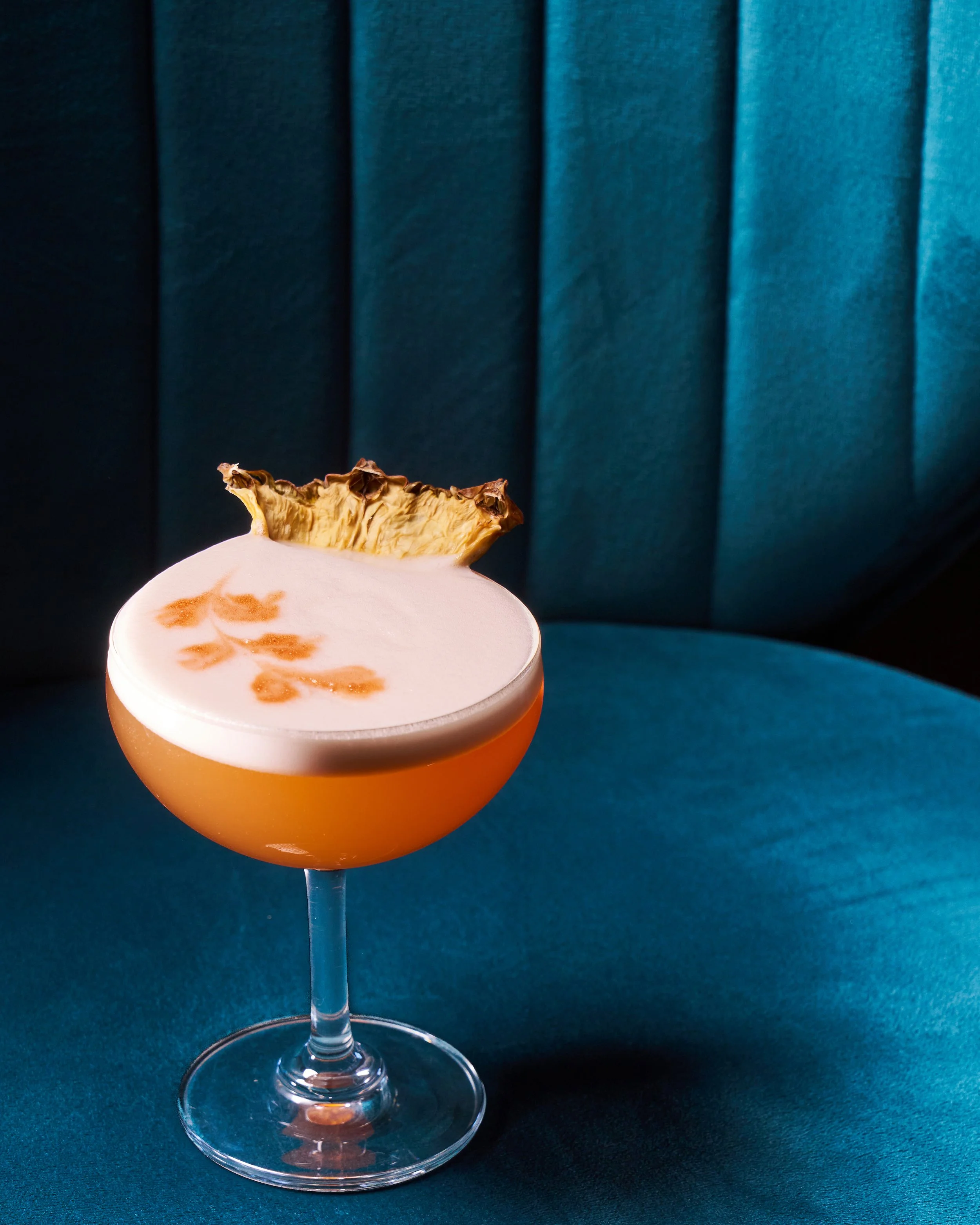A cocktail in a coupe glass with a frothy top, garnished with a dried pineapple slice on the rim, placed on a teal upholstered surface.