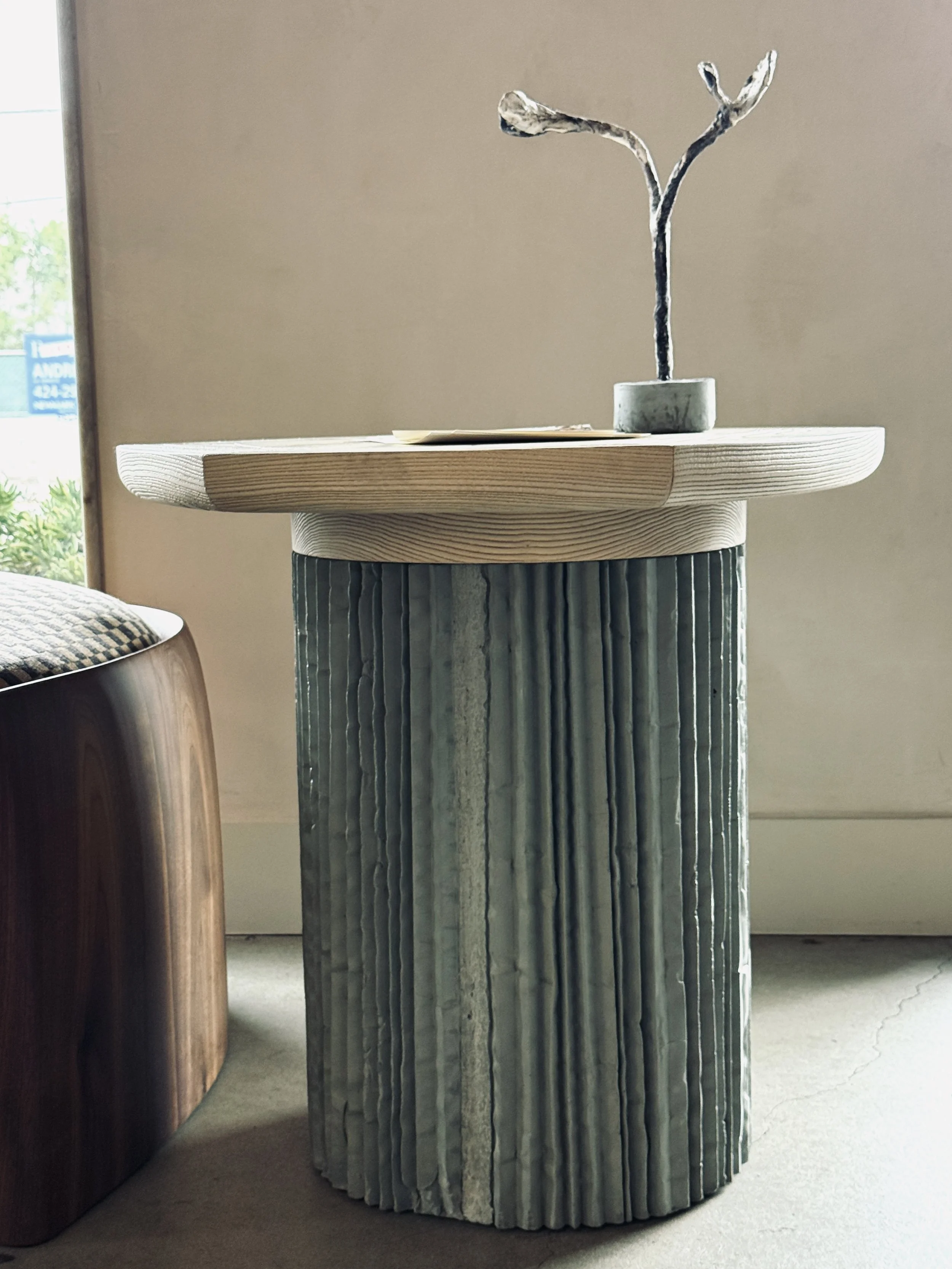 Plinth series of concrete table bases by O.M. Studio. Made from hand-scored pleating fabric paper. Side table in olive with Kept LA wax floral object on table -  at Stahl + Band, Los Angeles.