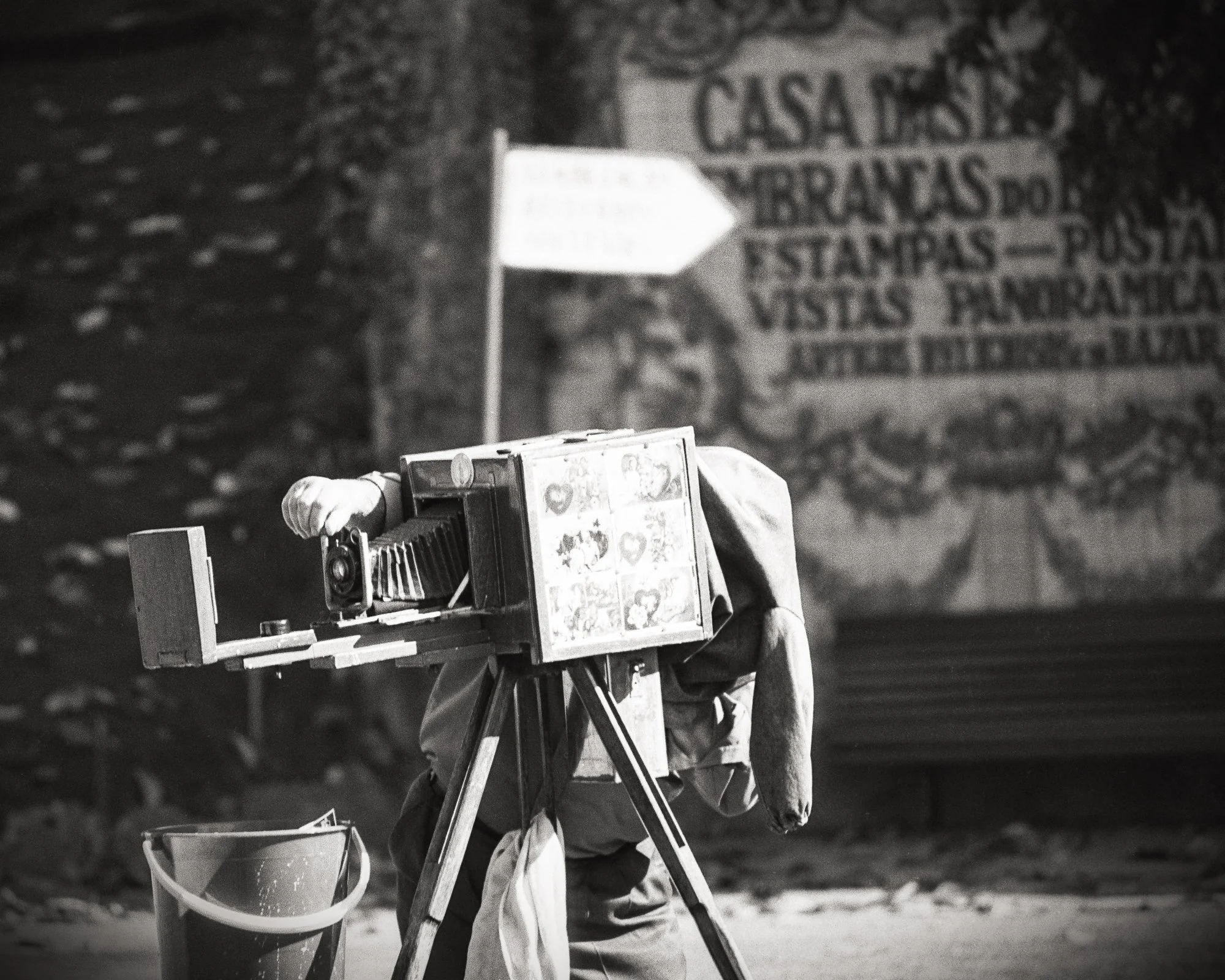 A vintage camera on a tripod, with a person adjusting it, stands on the street at night. In the background, a wall with Spanish text about a house with various services is partially visible.