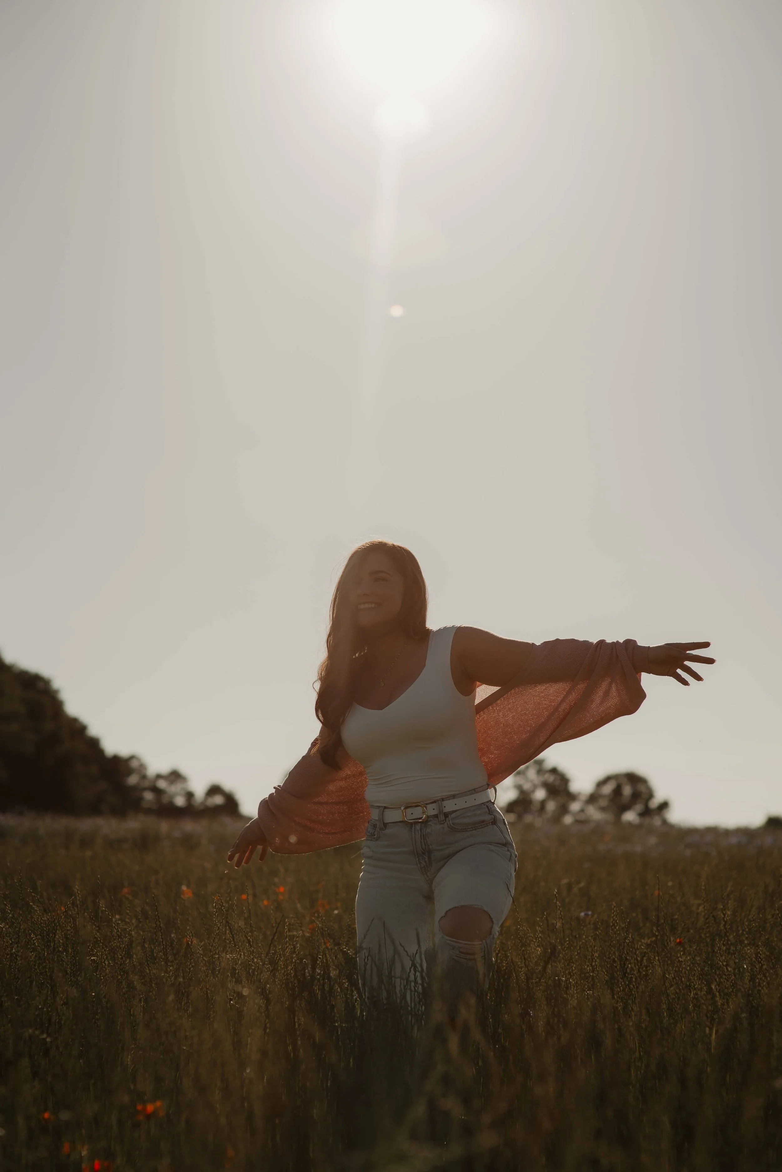 A woman standing in a field during sunset, smiling, with one arm extended outward, wearing a white tank top, denim shorts, and a pinkish-orange cardigan.