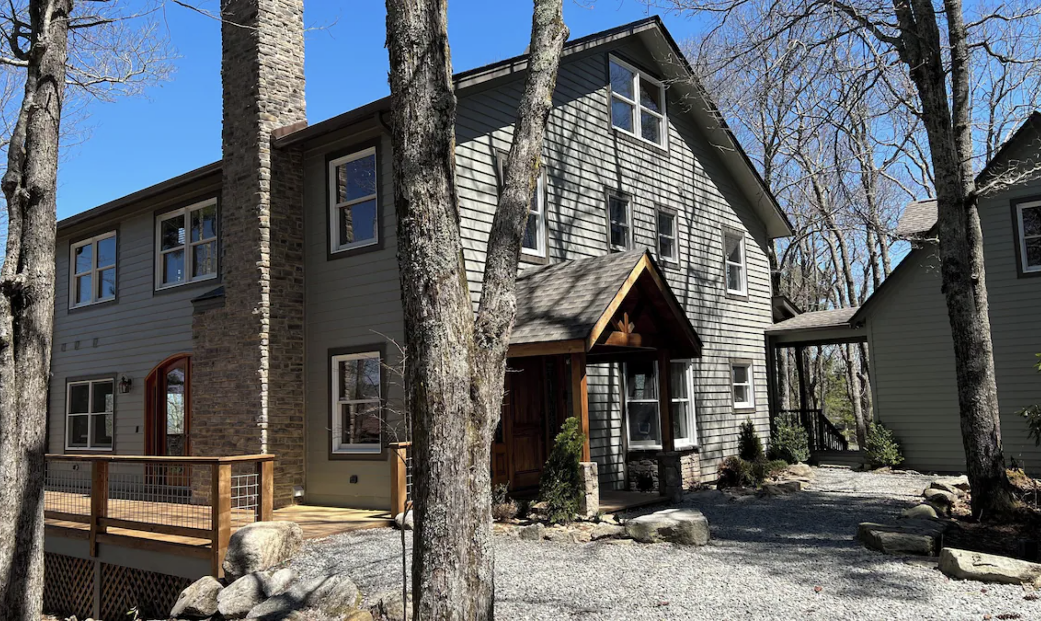 A multi-story house with gray siding, a stone chimney, and a small wooden porch, surrounded by leafless trees and a gravel yard under a clear blue sky.