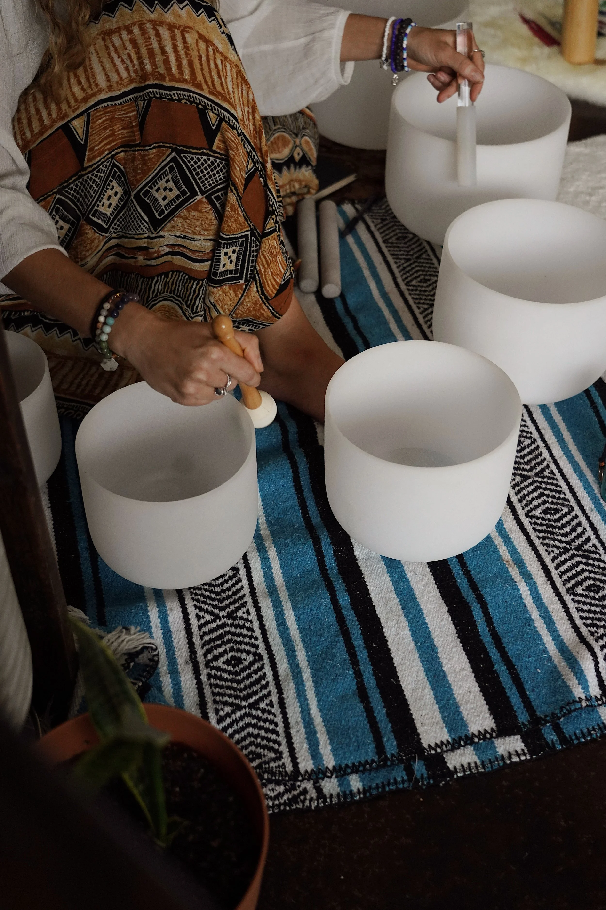 Person playing a singing bowl with a mallet, surrounded by various white singing bowls on a colorful woven cloth, with a potted plant and other objects in the background.