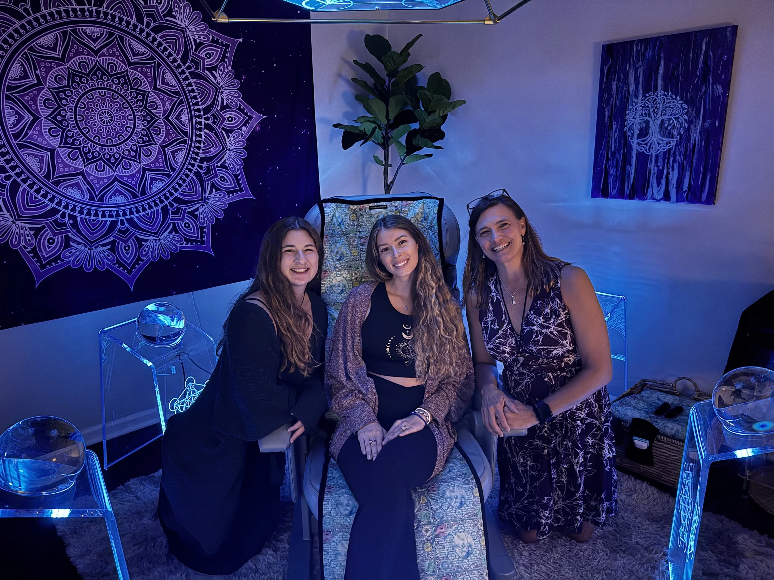 Three women smiling and posing for a photo indoors, with vibrant purple mandala and tree artwork on the wall behind them, in a room decorated with glowing blue acrylic furniture and plants.