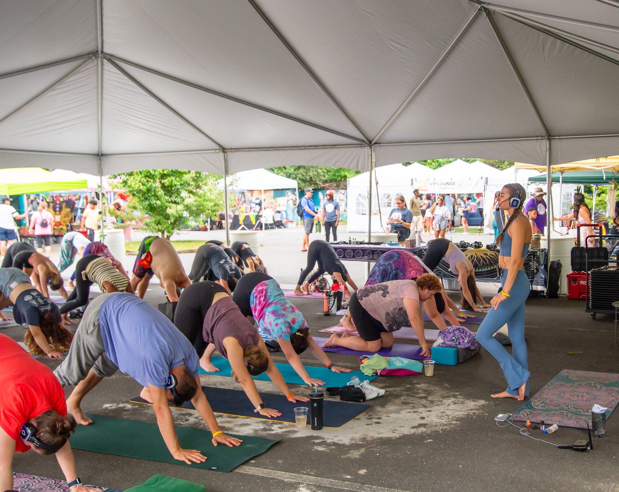 People participating in a yoga class under a large white canopy tent outdoors, with an instructor leading the session and other attendees in downward dog poses, surrounded by tents and booths at an outdoor event.