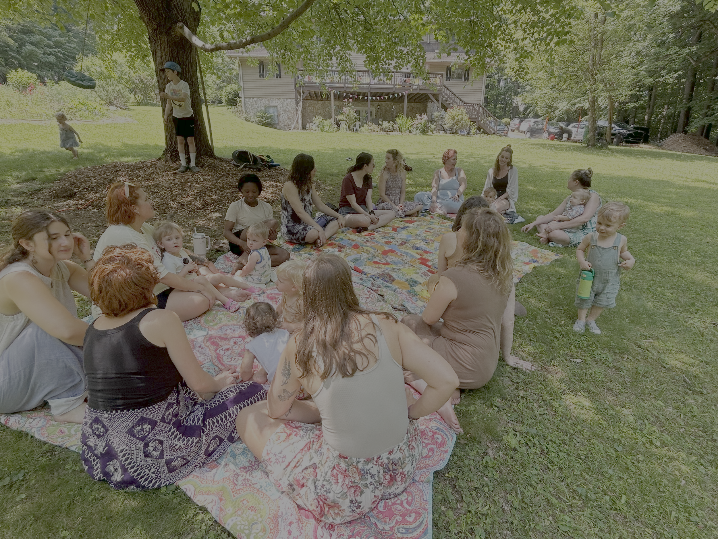 Group of women and children sitting on a blanket outdoors in a park, with a boy standing near a tree and a girl holding a water bottle.