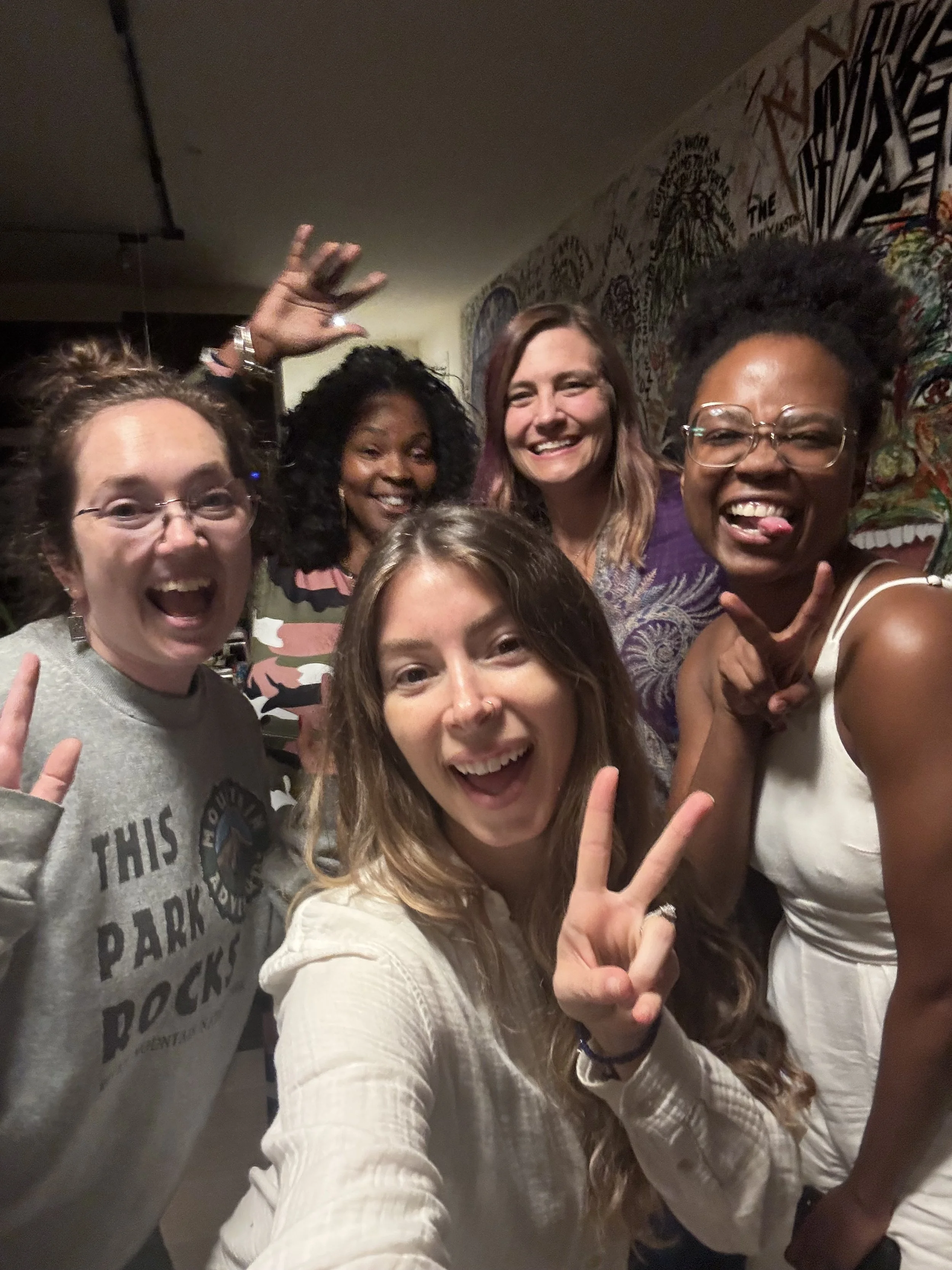 Group of five women smiling and making playful gestures in front of colorful graffiti wall.