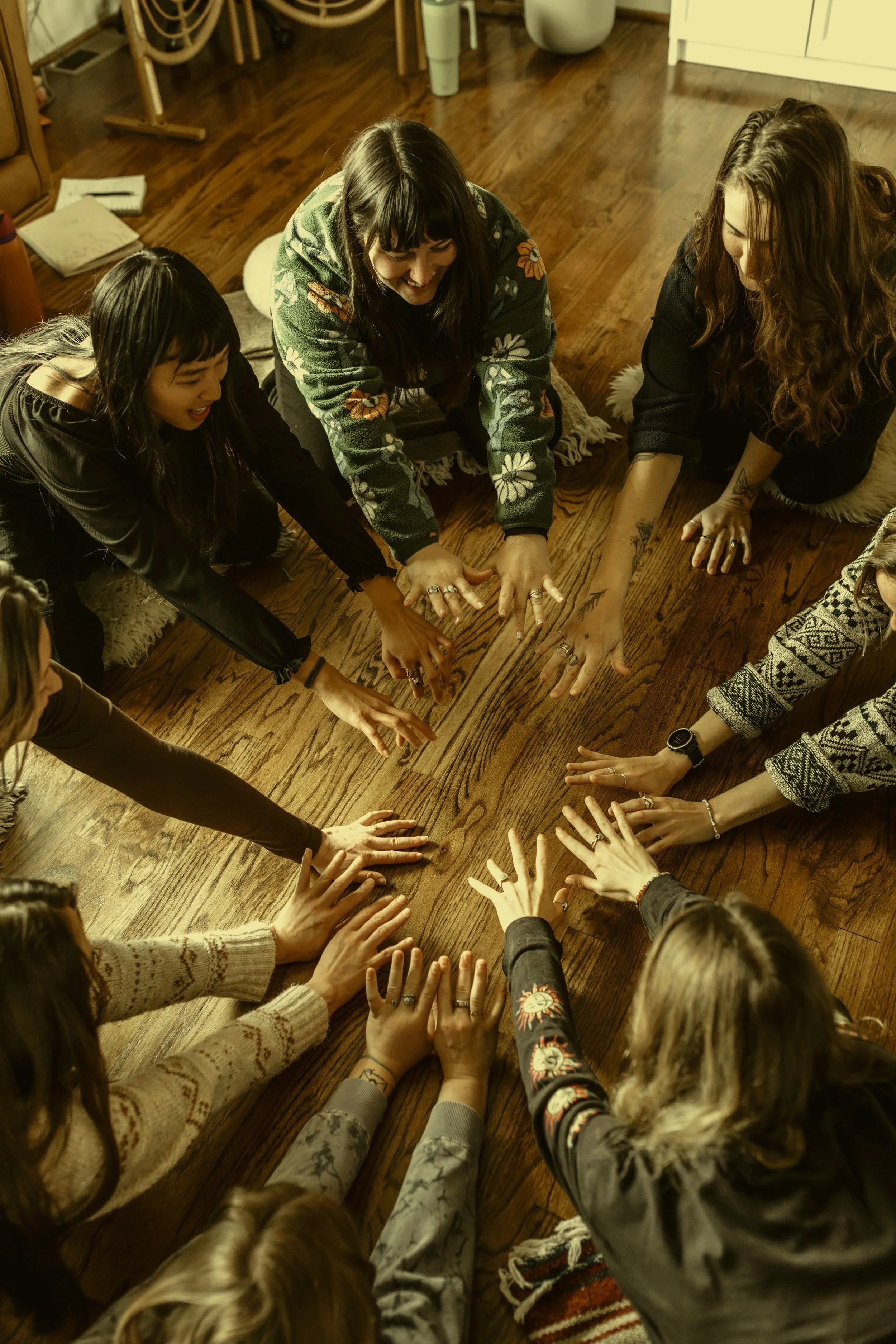 A group of diverse women and girls sitting on the wooden floor, reaching out and touching the center of a wooden table together, smiling and engaging in a team activity.