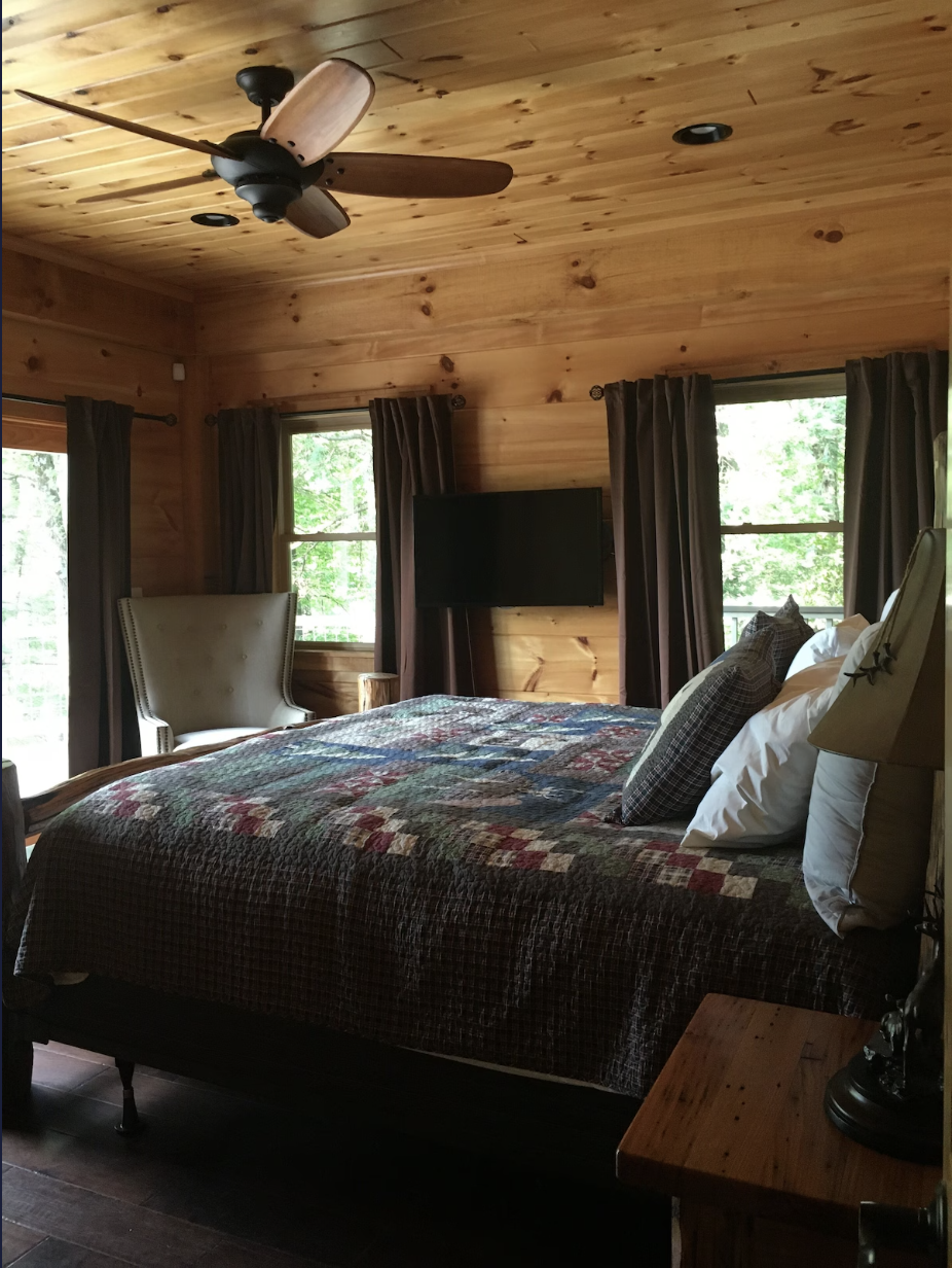 A cozy bedroom with wooden walls and ceiling, featuring a bed with a quilt, pillows, a bedside table with a lamp, a chair, a wall-mounted TV, and three windows with dark curtains showing greenery outside.