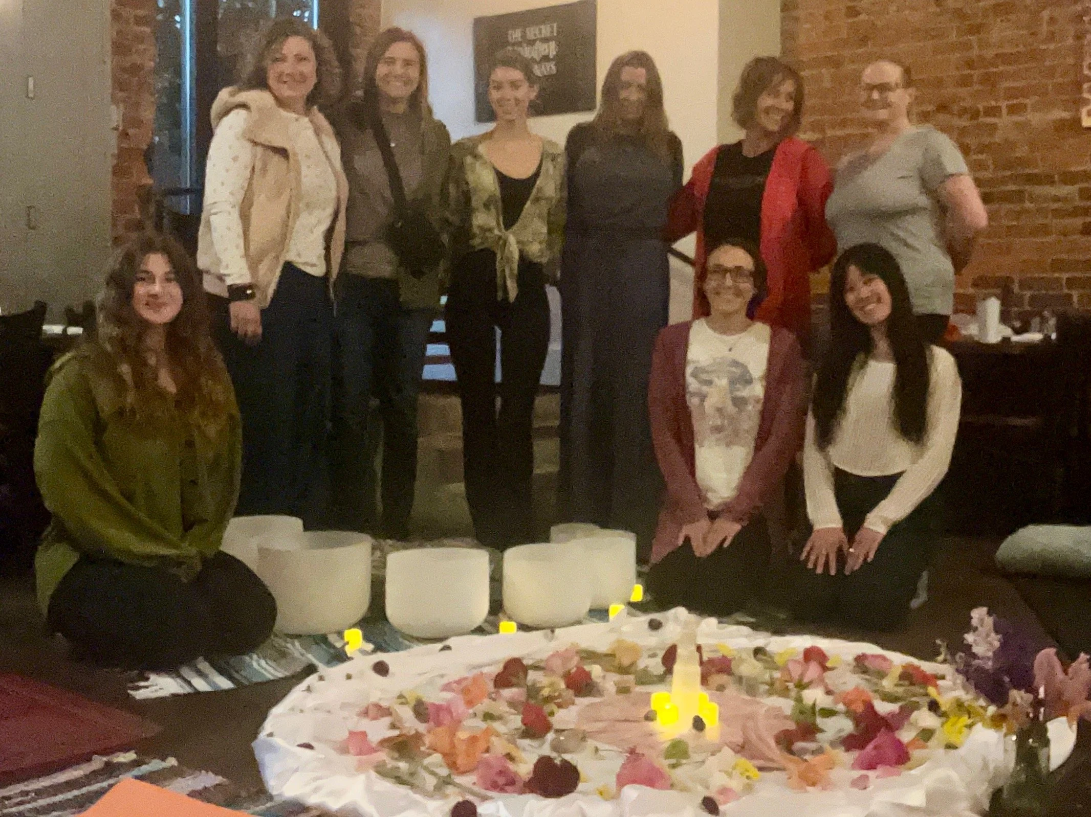 A group of nine women, some kneeling and some standing, celebrating around a large flower petal-covered cake with candles, in a cozy brick-walled setting.