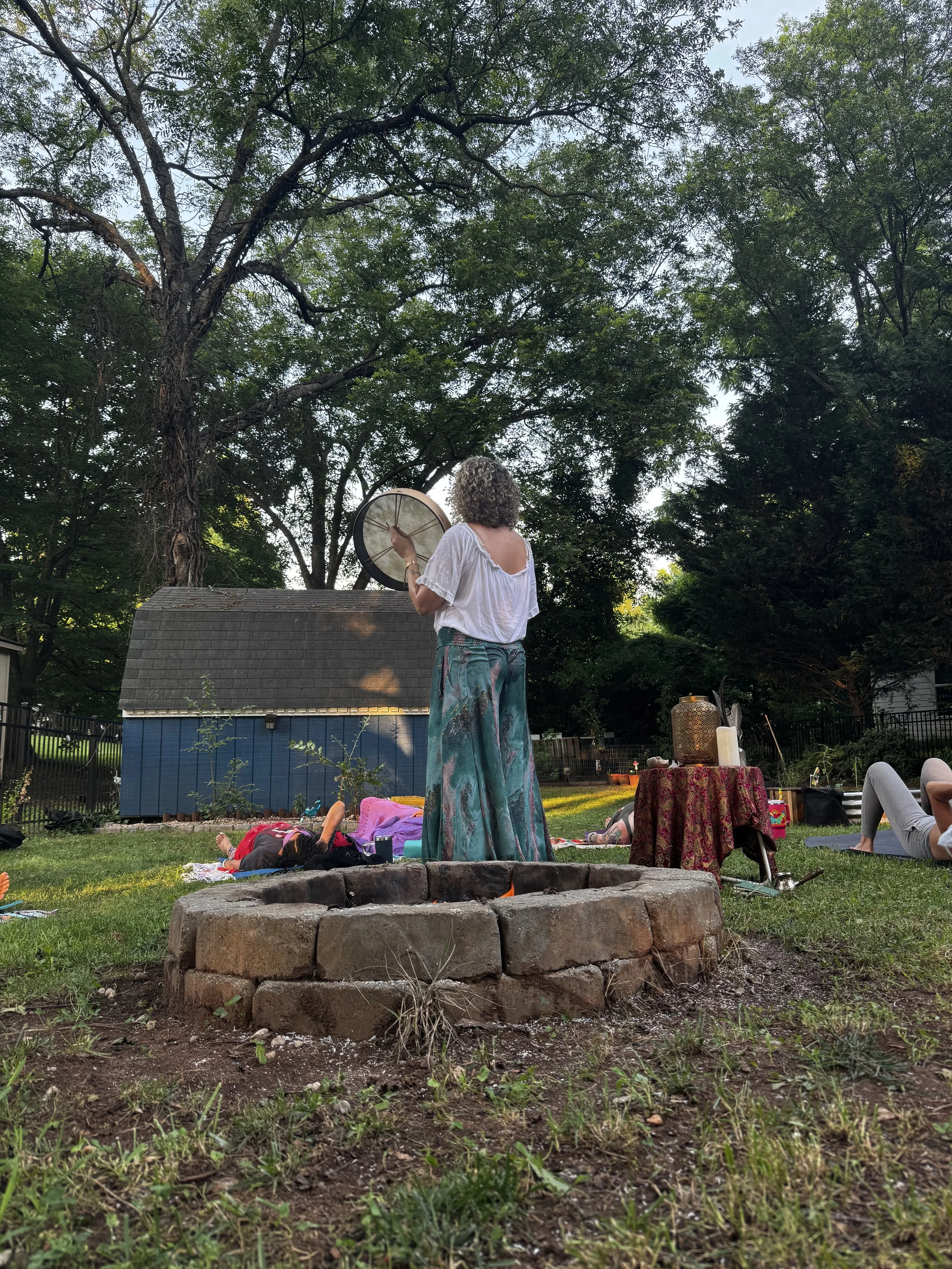 A woman holding a drum standing on grass near a fire pit, with people lying on blankets and a table with candles nearby, in a backyard with trees.
