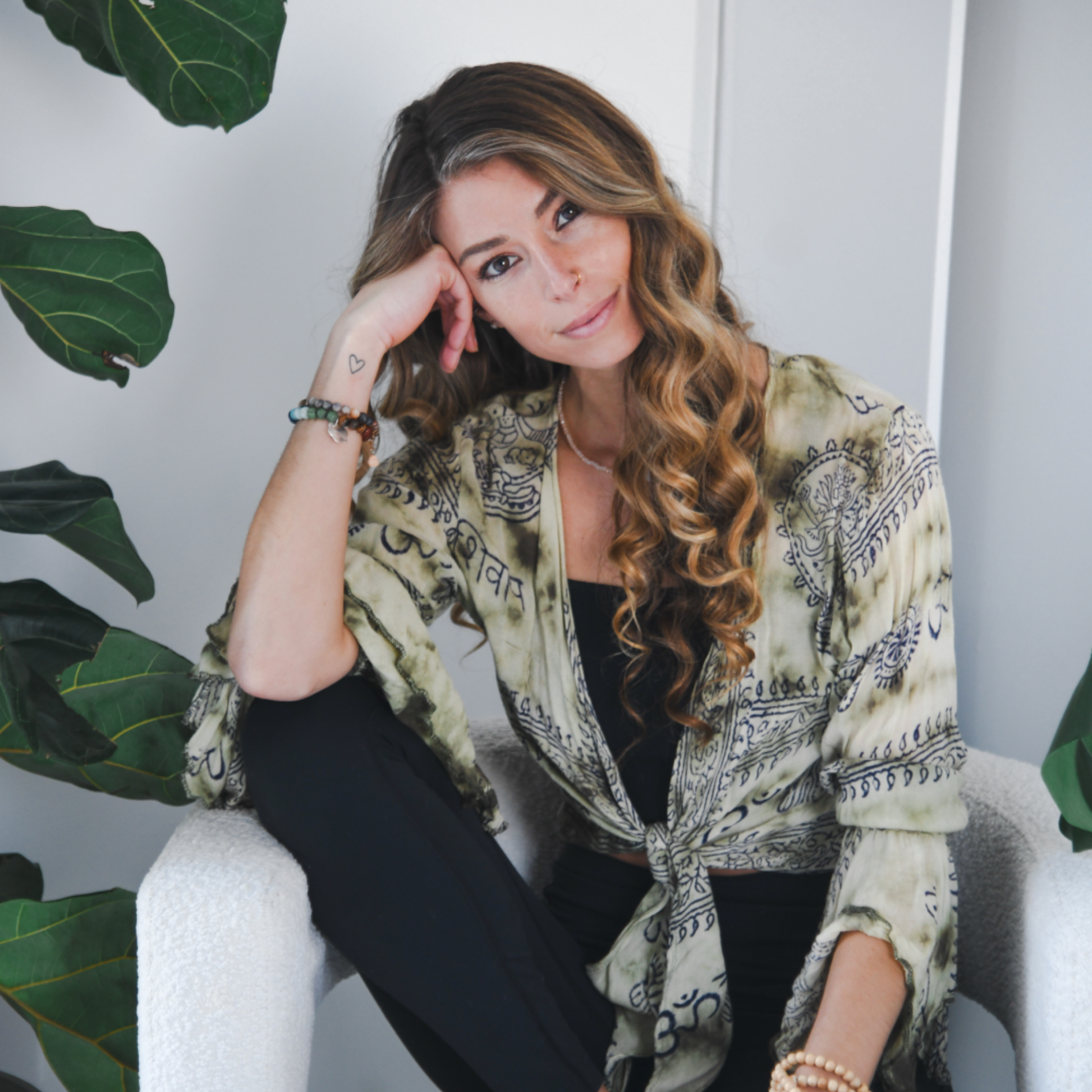 A woman with long curly hair sitting on a white chair, resting her head on her hand, surrounded by green plants, wearing a patterned beige shirt and black pants.