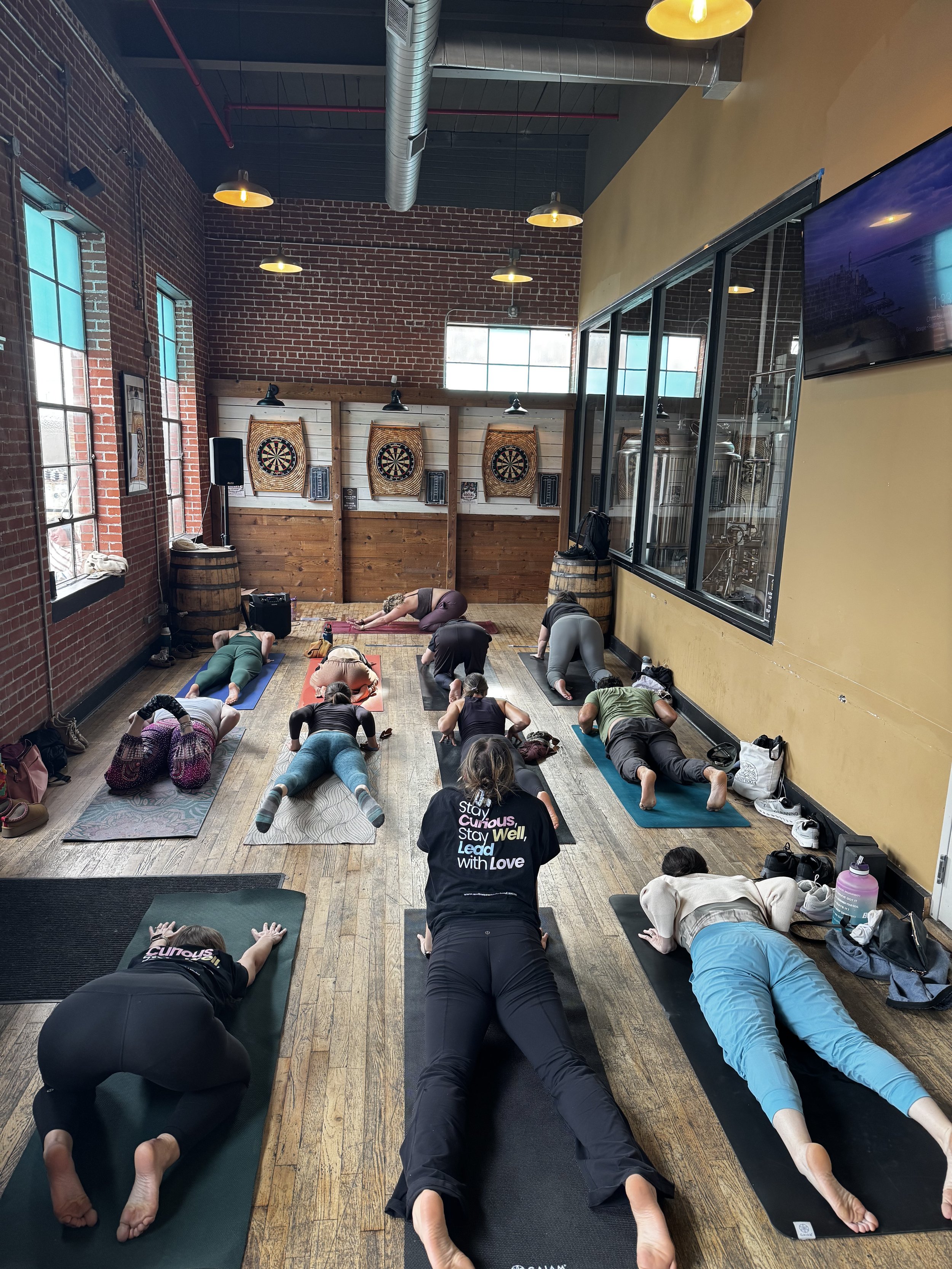 People participating in a yoga class in a rustic studio with exposed brick walls, large windows, and protruding ductwork. Participants are on yoga mats in various poses, guided by an instructor at the front.