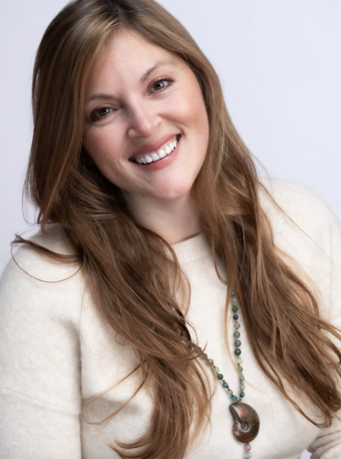 A woman with long, wavy red hair smiling and looking at the camera, wearing a beige sweater and a beaded necklace with a large pendant.