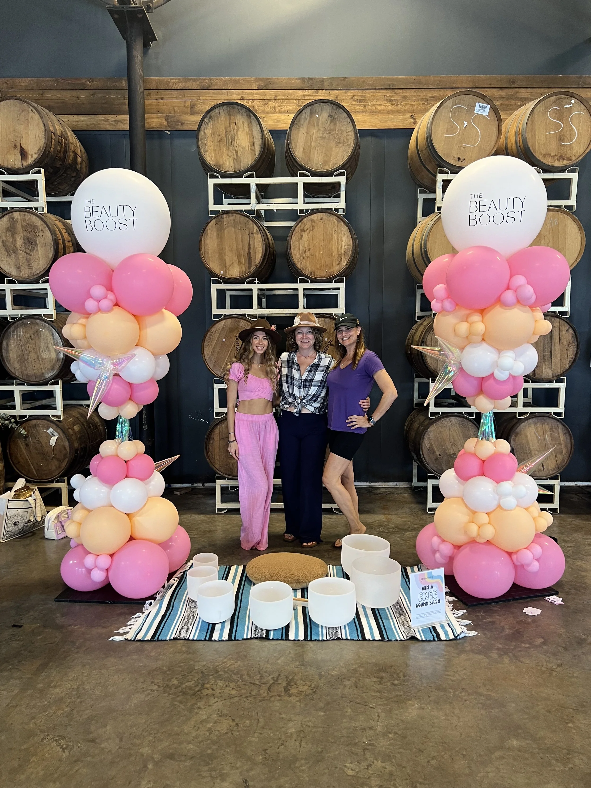 Three women standing in front of large whiskey barrels with pink and peach balloon columns on each side. The woman in the middle wears a checkered shirt and a hat, the woman on the left wears a pink outfit, and the woman on the right wears a purple s