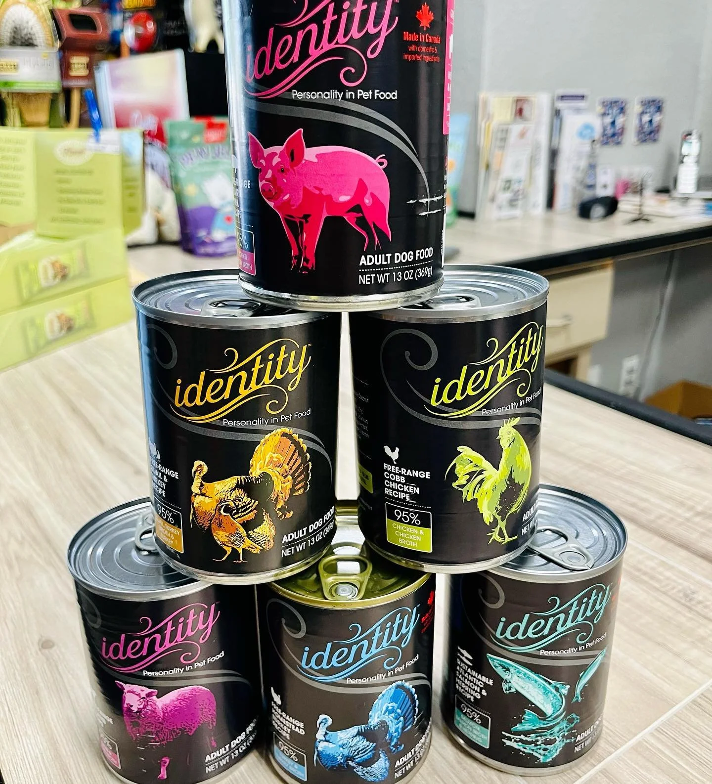Six cans of Identity brand pet food arranged in a pyramid on a wooden surface. The cans feature colorful animal illustrations and text indicating flavors like turkey, chicken, and salmon, with the top can showing a pink pig.